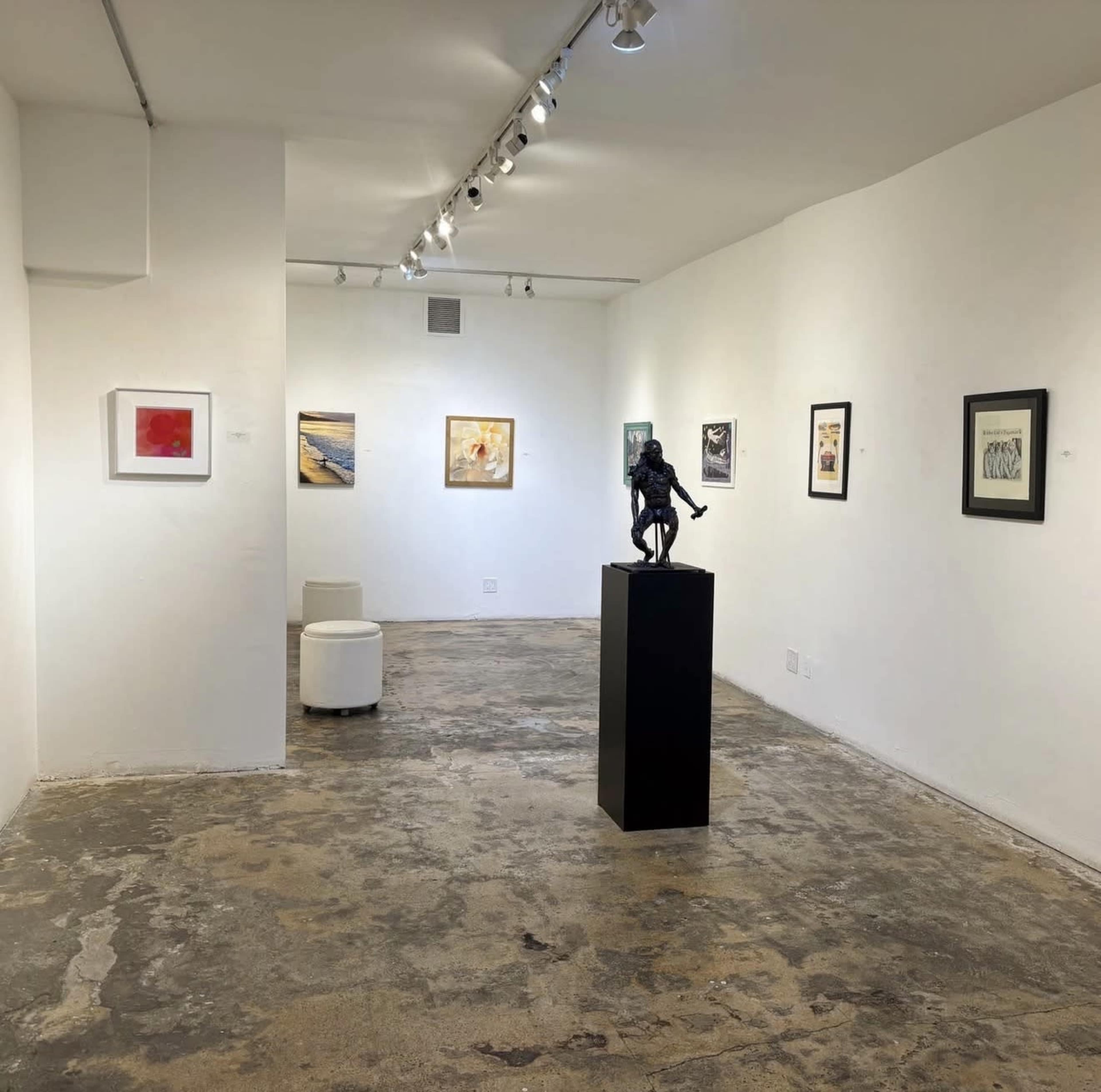 The image depicts a contemporary art gallery with several framed artworks on the walls and a sculpture displayed on a pedestal in the center.