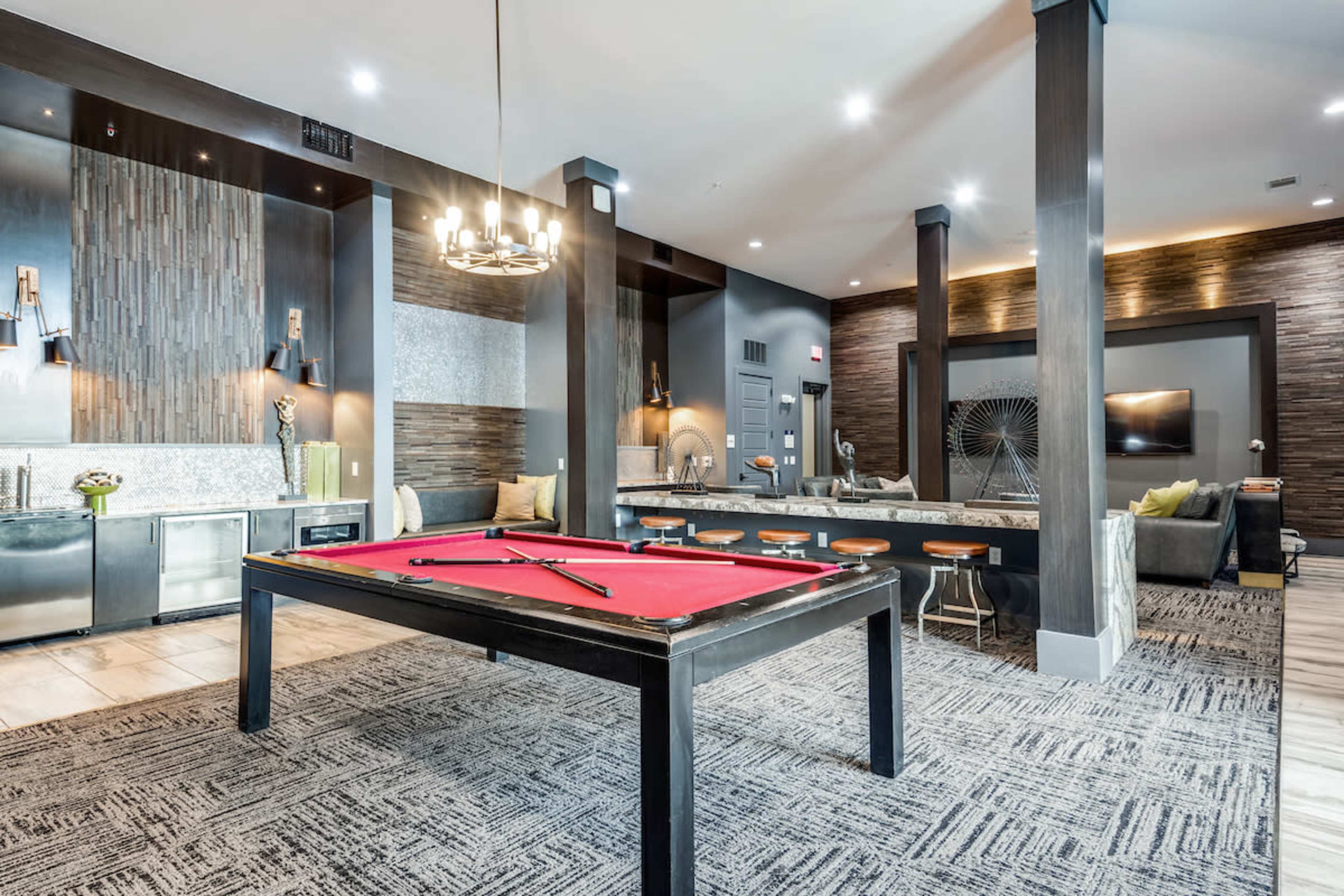 A modern lounge area with a red pool table, bar stools, and contemporary furniture set against a stylish backdrop of textured wall panels and pendant lighting.