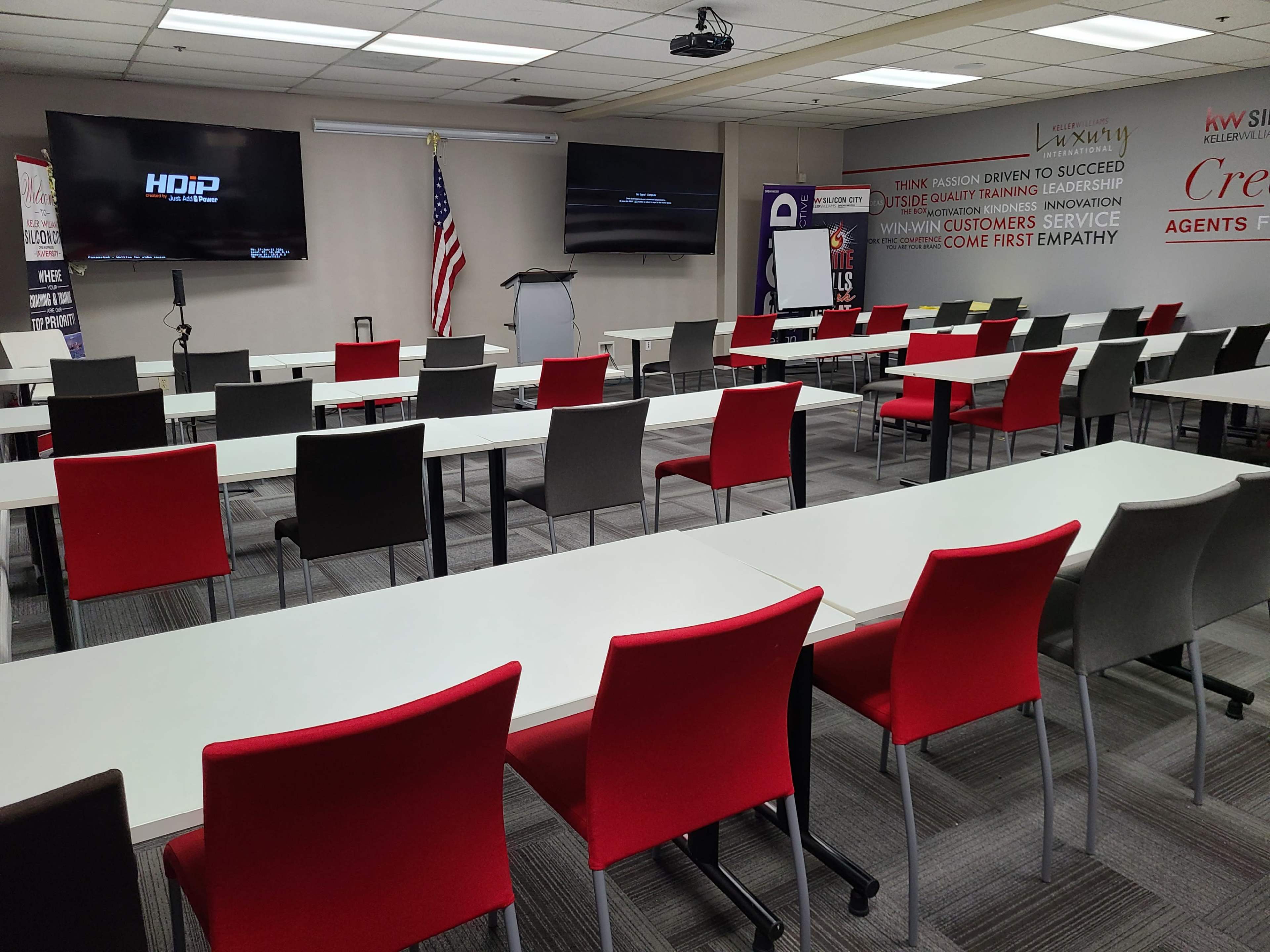A classroom setting features arranged tables and chairs in red and gray, with an American flag and screens at the front.