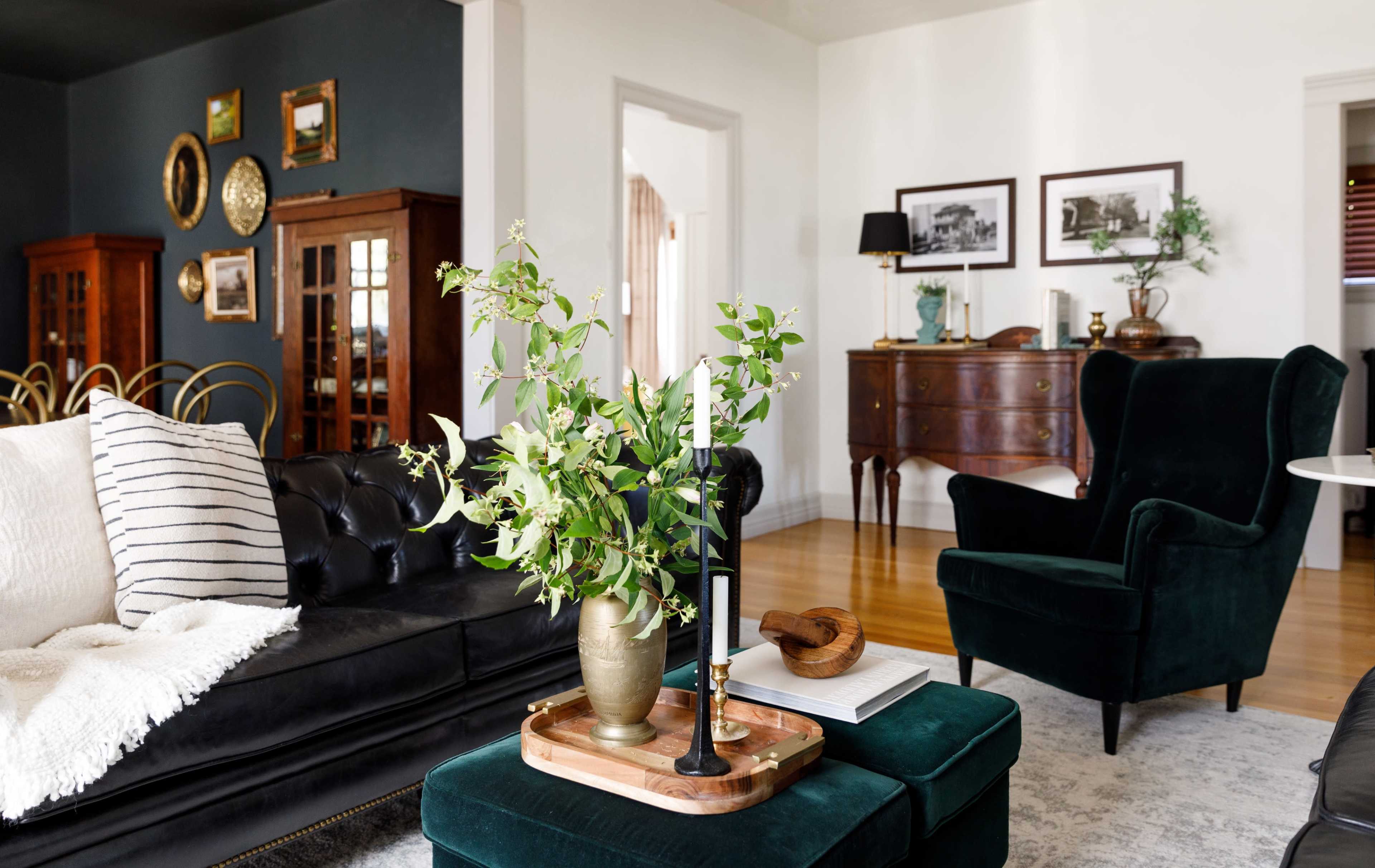 The living room features dark walls and a mix of vintage furniture, including a tufted black sofa, green armchairs, and a wooden sideboard, complemented by decorative plants and framed photographs.