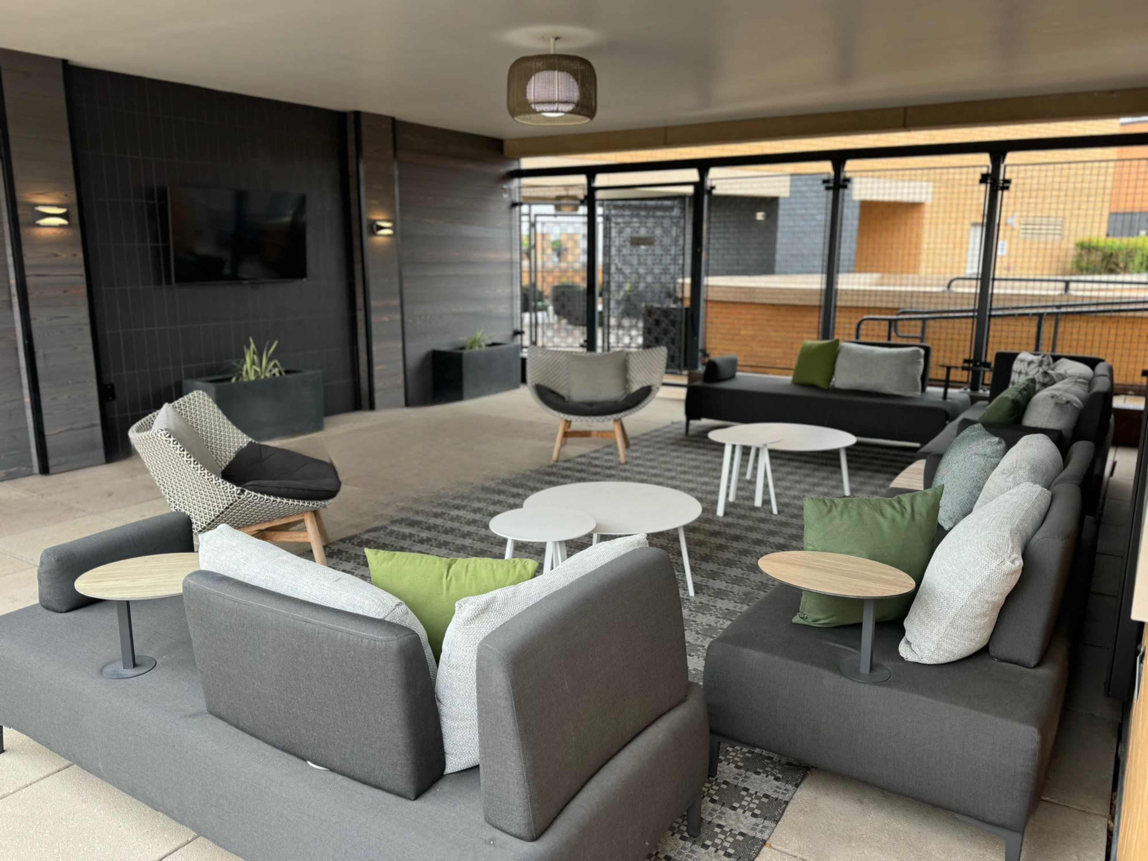 A modern lounge area with multiple gray sofas, round tables, and green accent pillows, enclosed by a glass wall.