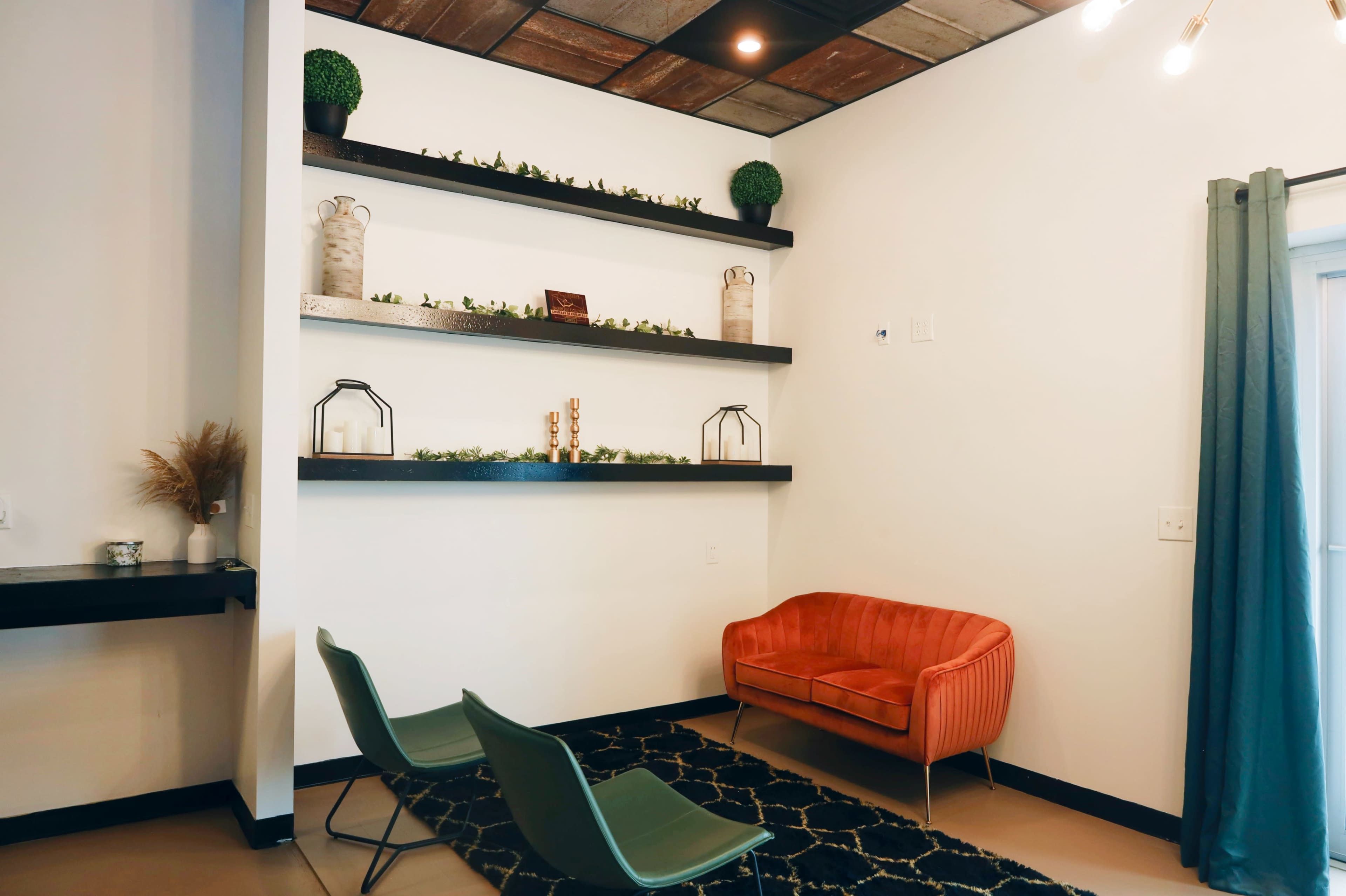 A small seating area with a coral-colored couch and two green chairs beneath a wall featuring floating shelves adorned with decorative items.