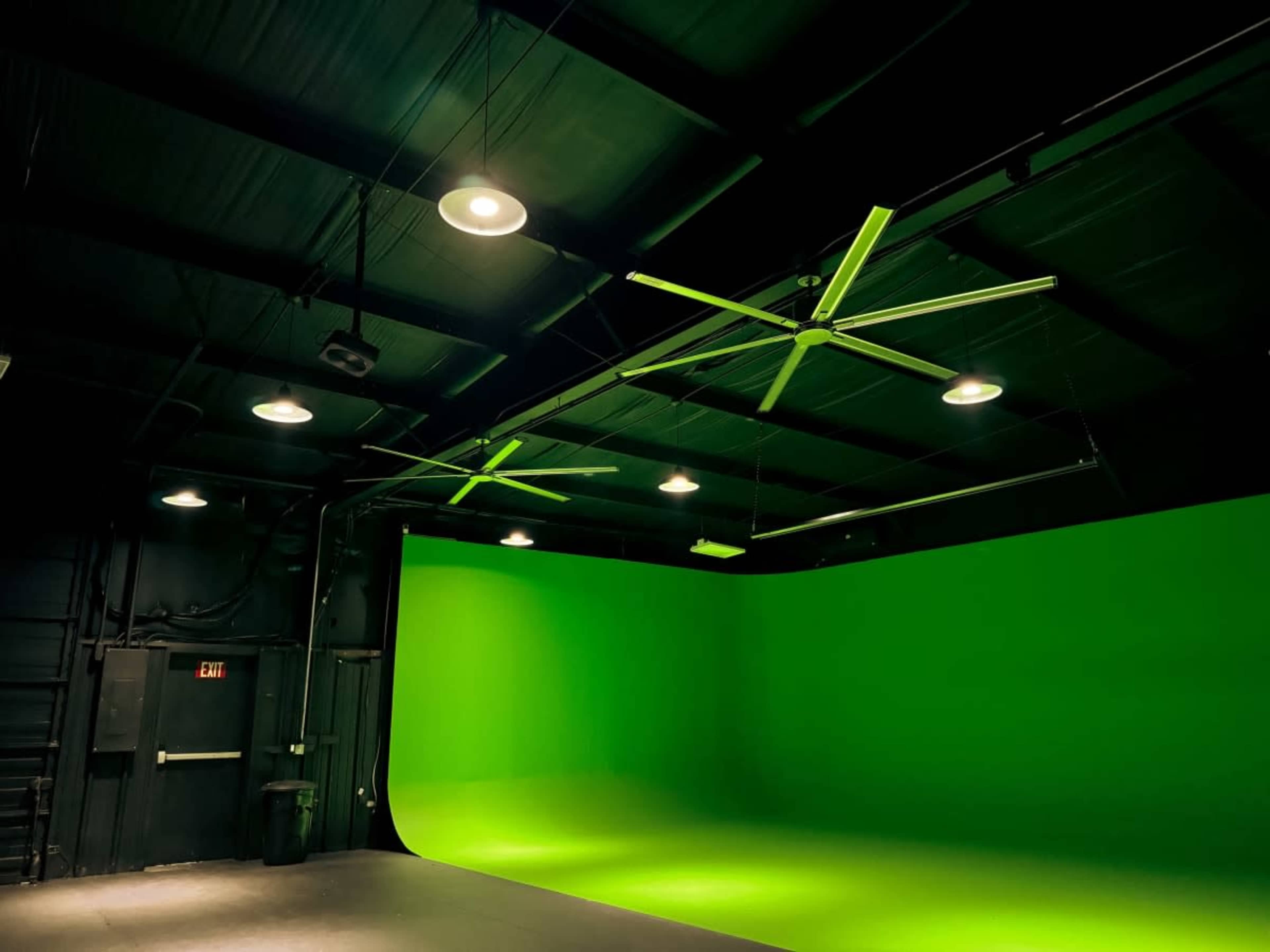 The image shows a studio space with a green screen backdrop and large ceiling fans.