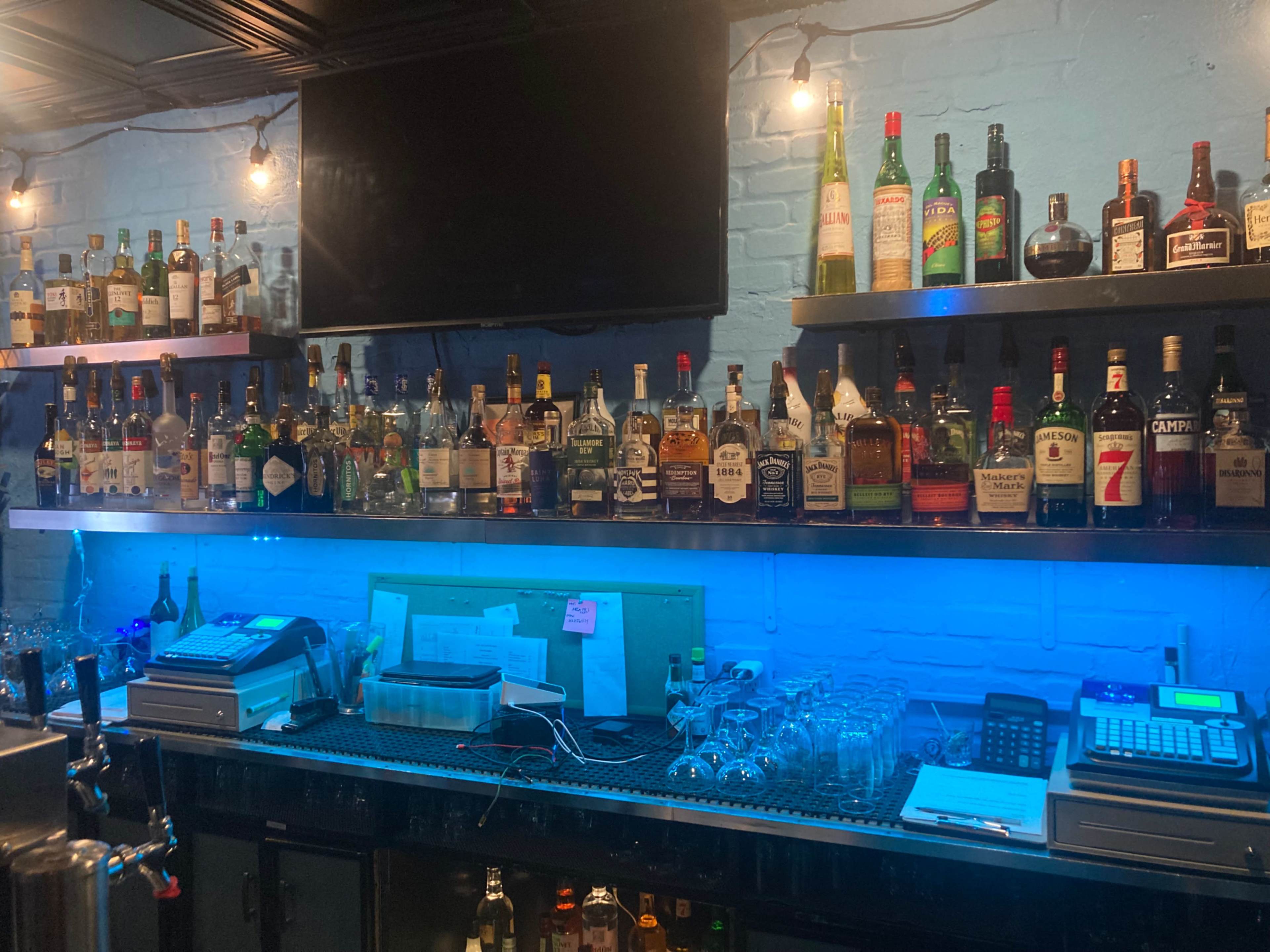 The image shows a bar with shelves lined with various bottles of liquor above a bar counter equipped with a cash register and glasses.
