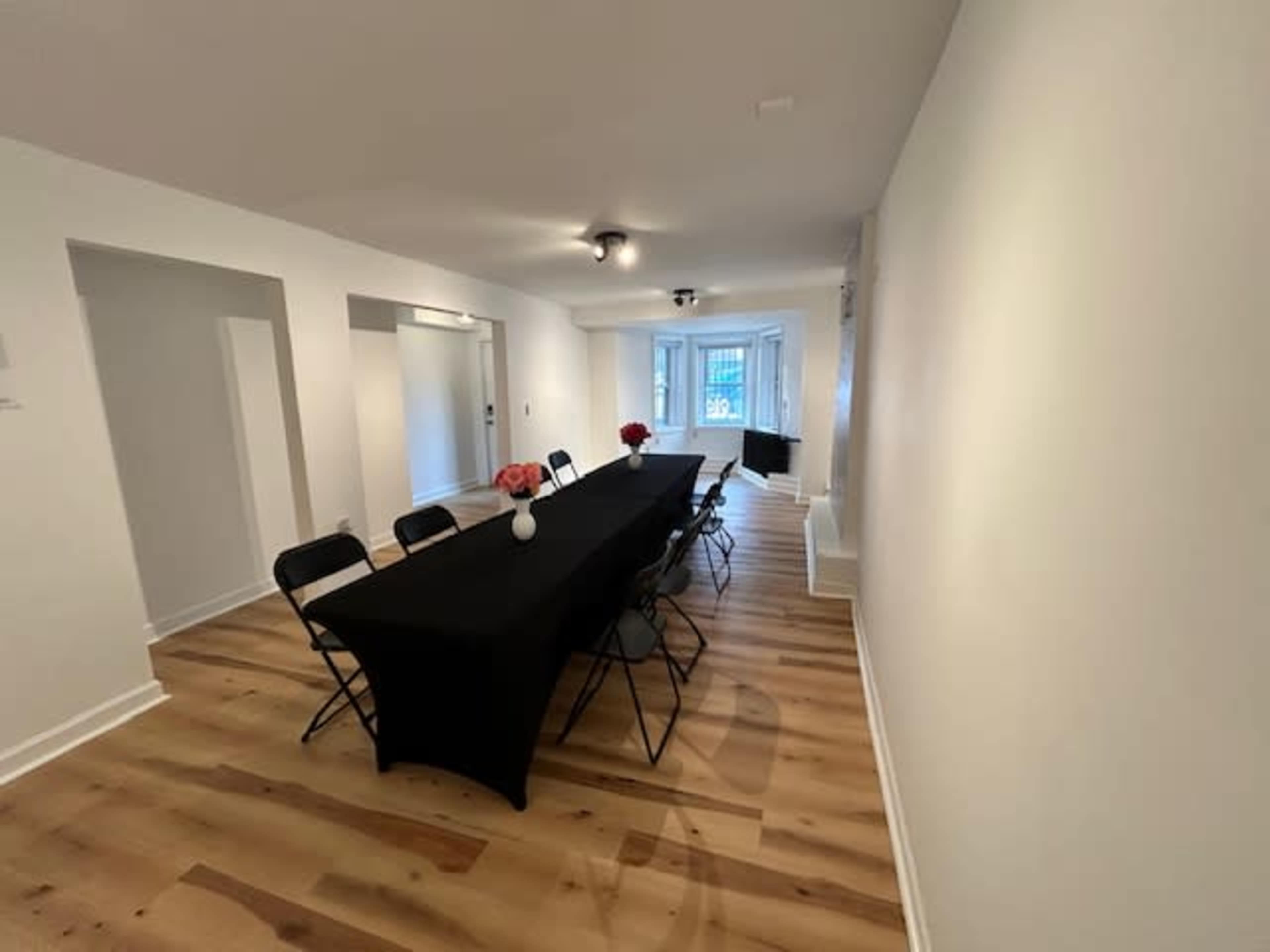 A long black table with chairs set in a bright, minimalist room featuring wooden flooring and a window at the far end.