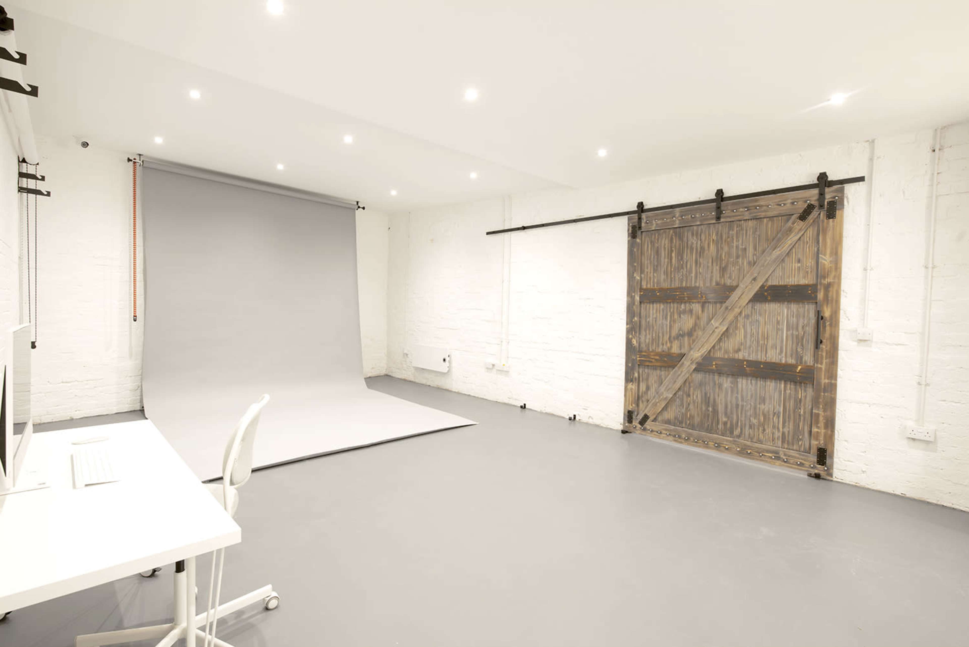 A spacious studio with a gray backdrop, wooden barn-style doors, and a minimalist white desk and chair.