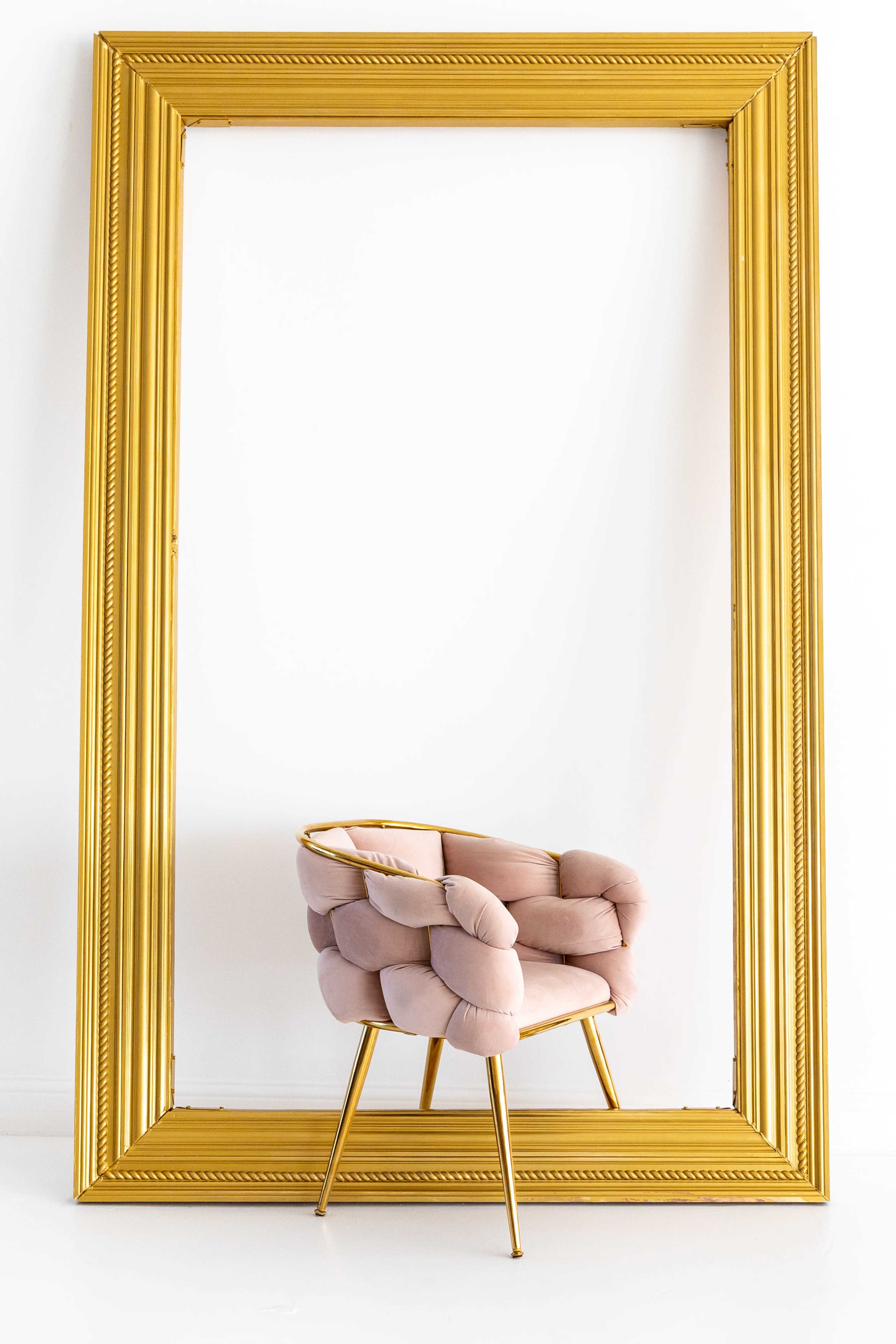 A plush pink chair is positioned in front of a large, ornate gold picture frame against a white wall.