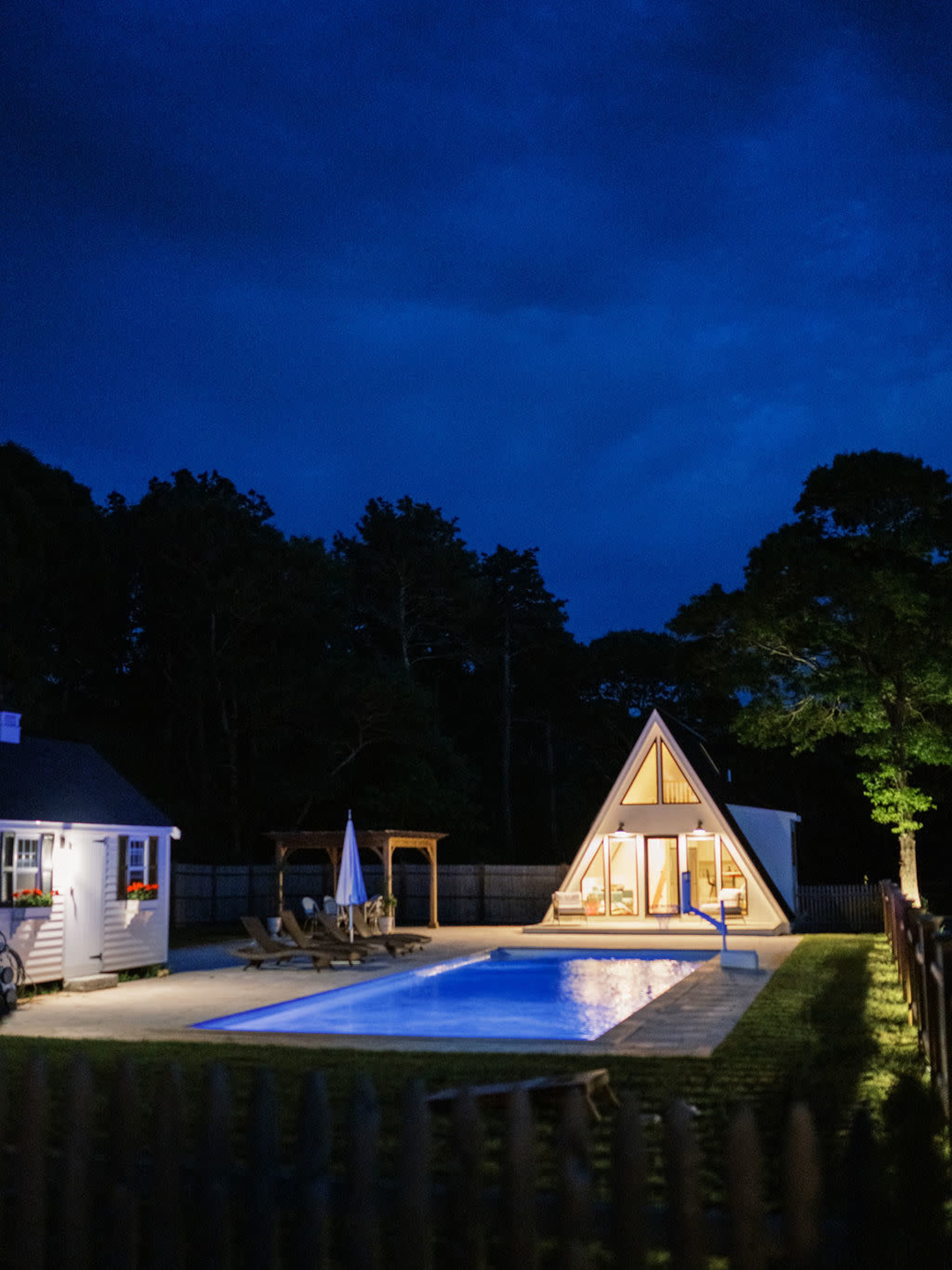 A triangular-roofed house with large windows and a pool illuminated at night, surrounded by trees and a fence.