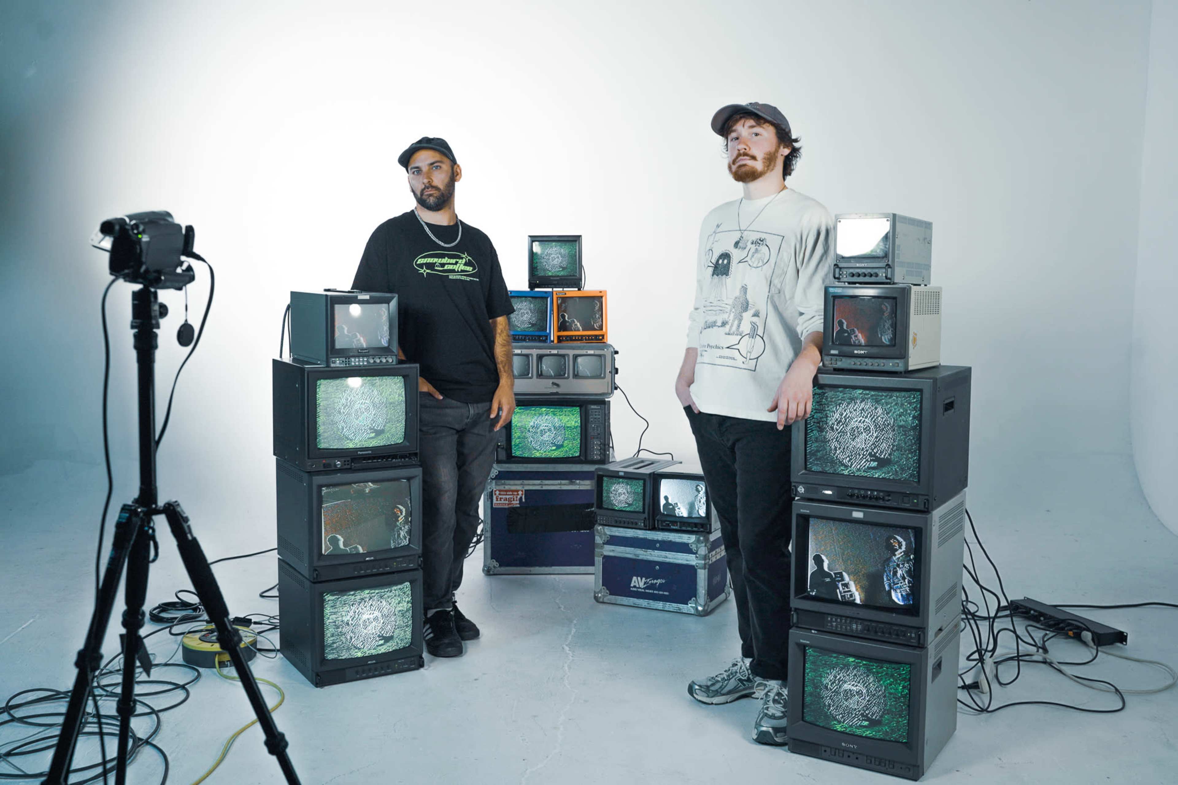 Two individuals stand next to a stack of vintage televisions in a studio setting, while a camera on a tripod captures the scene.