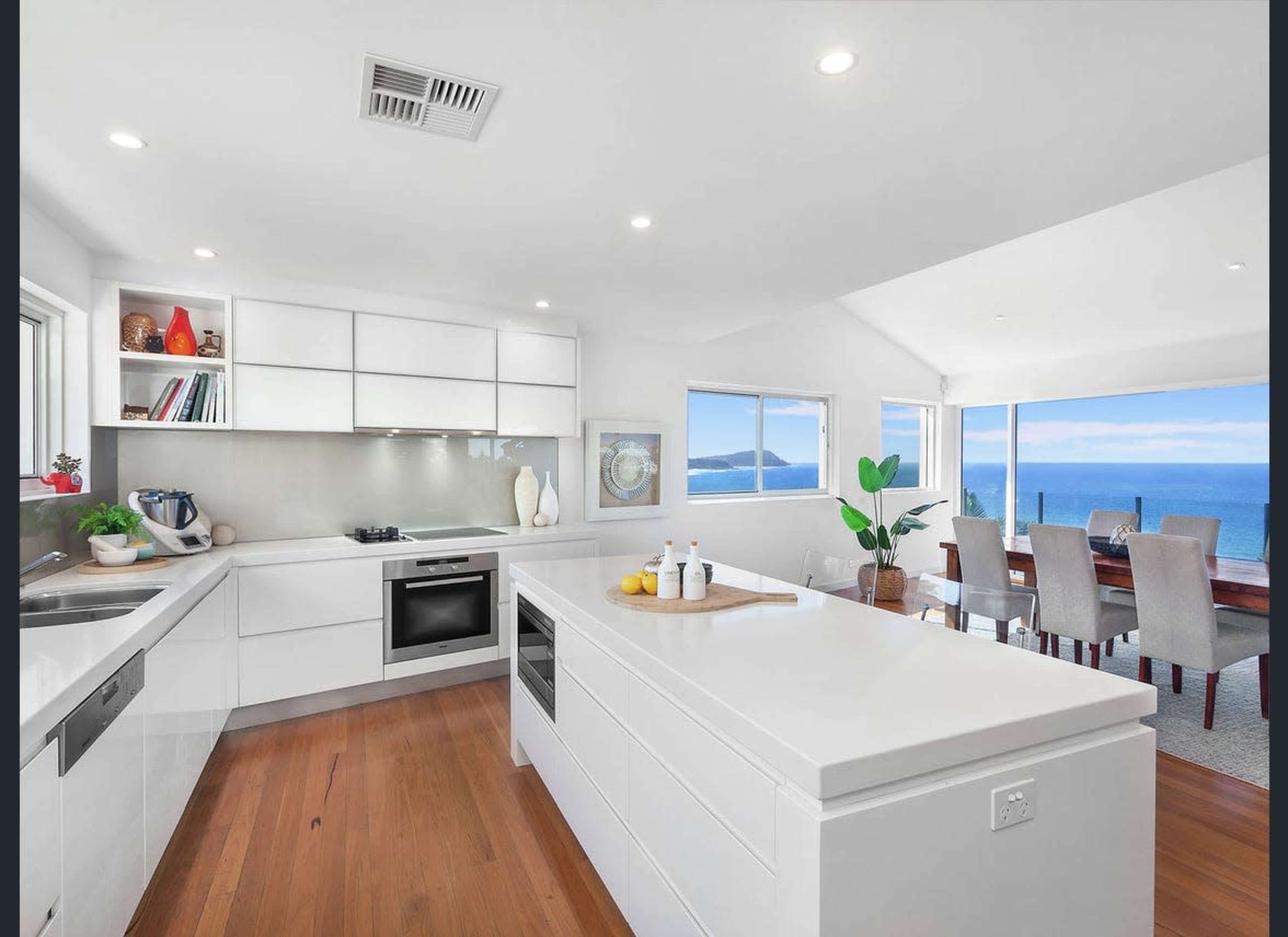 The image shows a modern kitchen with white cabinetry, a central island, and large windows overlooking a dining area with ocean views.