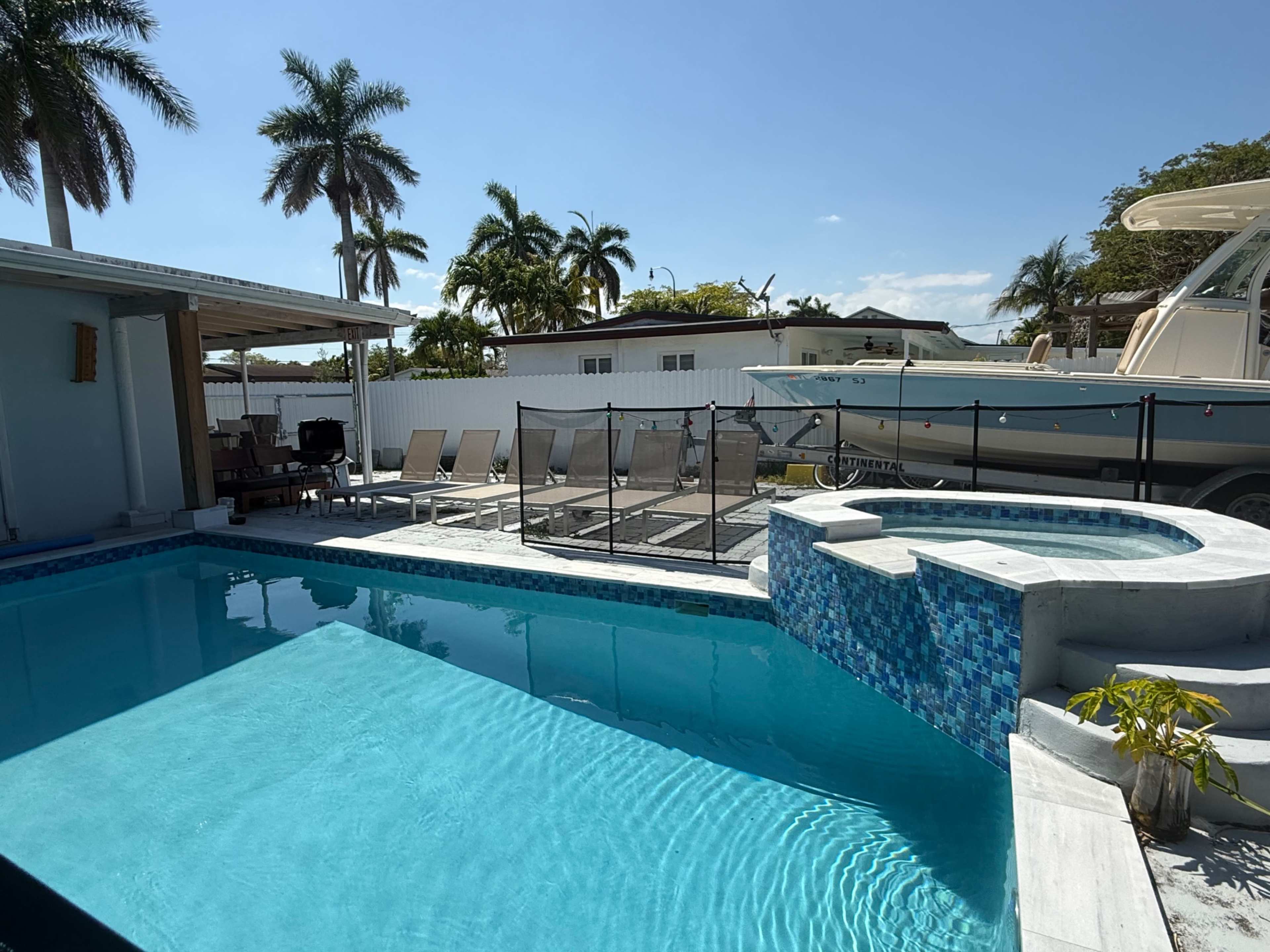 The image depicts a swimming pool area with a hot tub, surrounded by palm trees and lounge chairs, alongside a boat parked nearby.