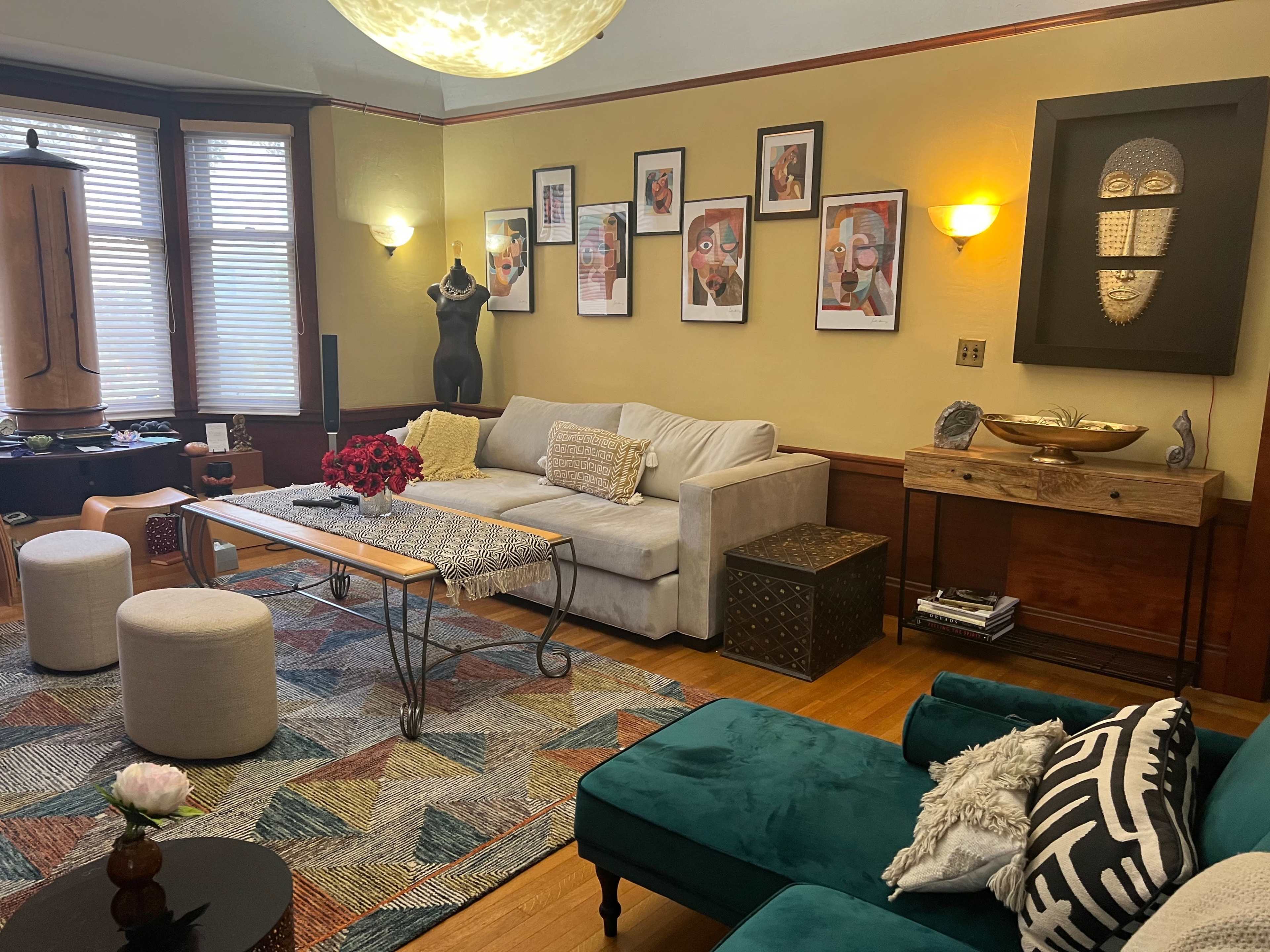 The image shows a cozy living room with a gray sofa, colorful artwork on the walls, a wooden coffee table, a plush green chair, and decorative accents.