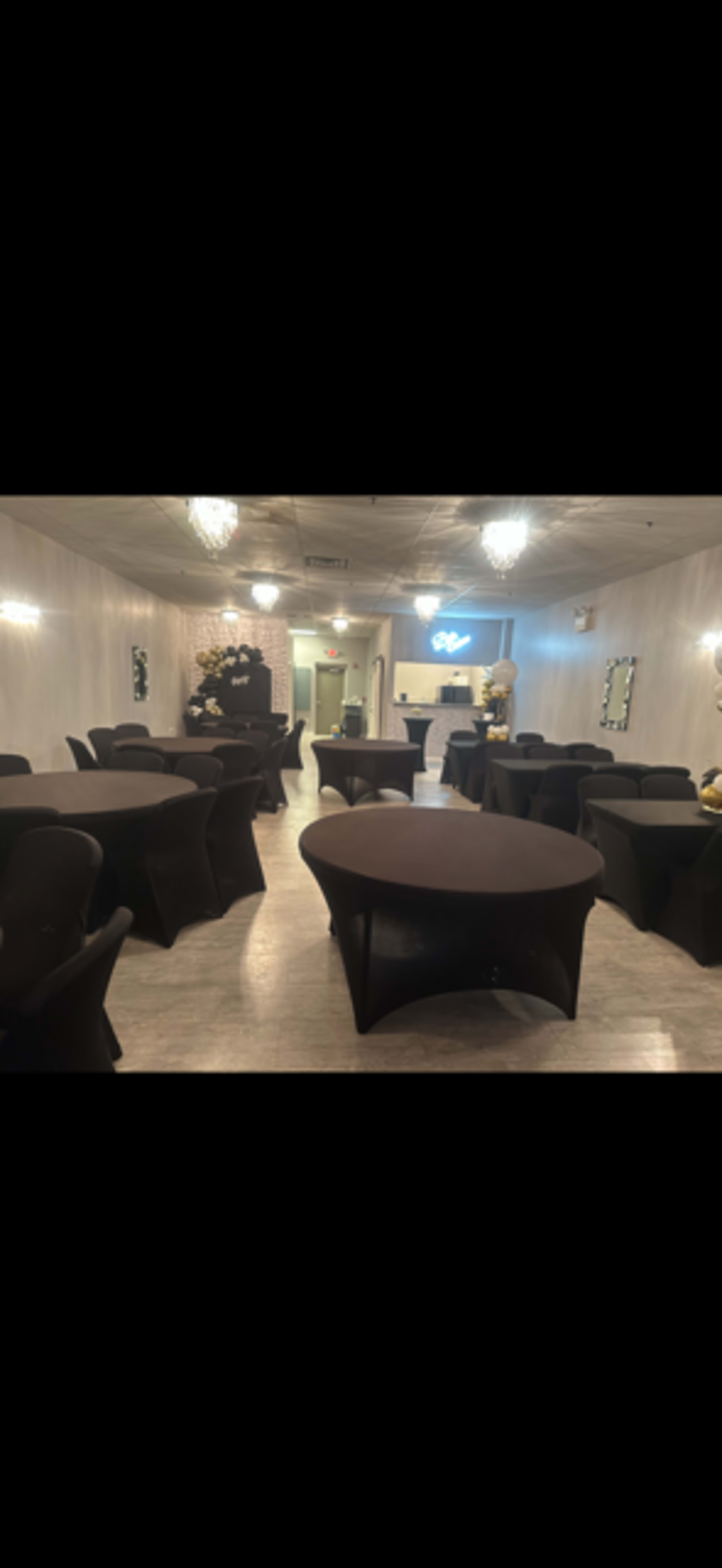 An elegantly arranged event space with round tables covered in black tablecloths and soft lighting from chandeliers above.