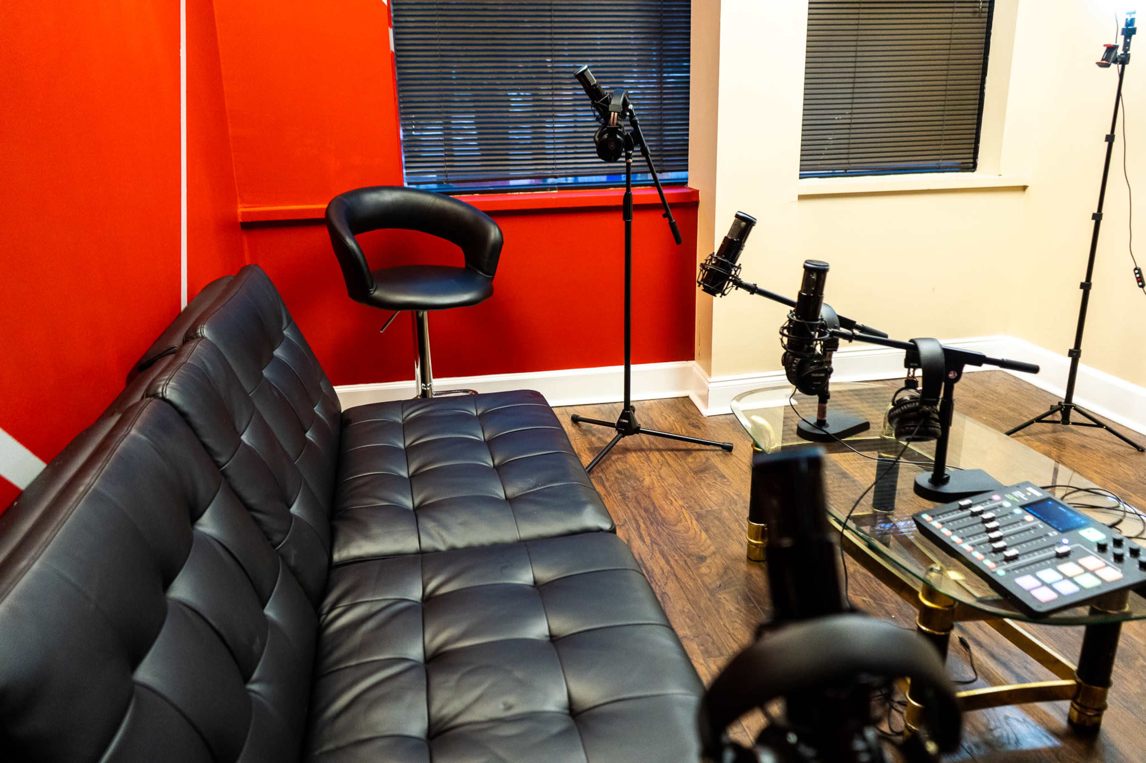 The image shows a recording studio with a black leather couch, a glass table, and multiple microphones on stands against a bright red wall.