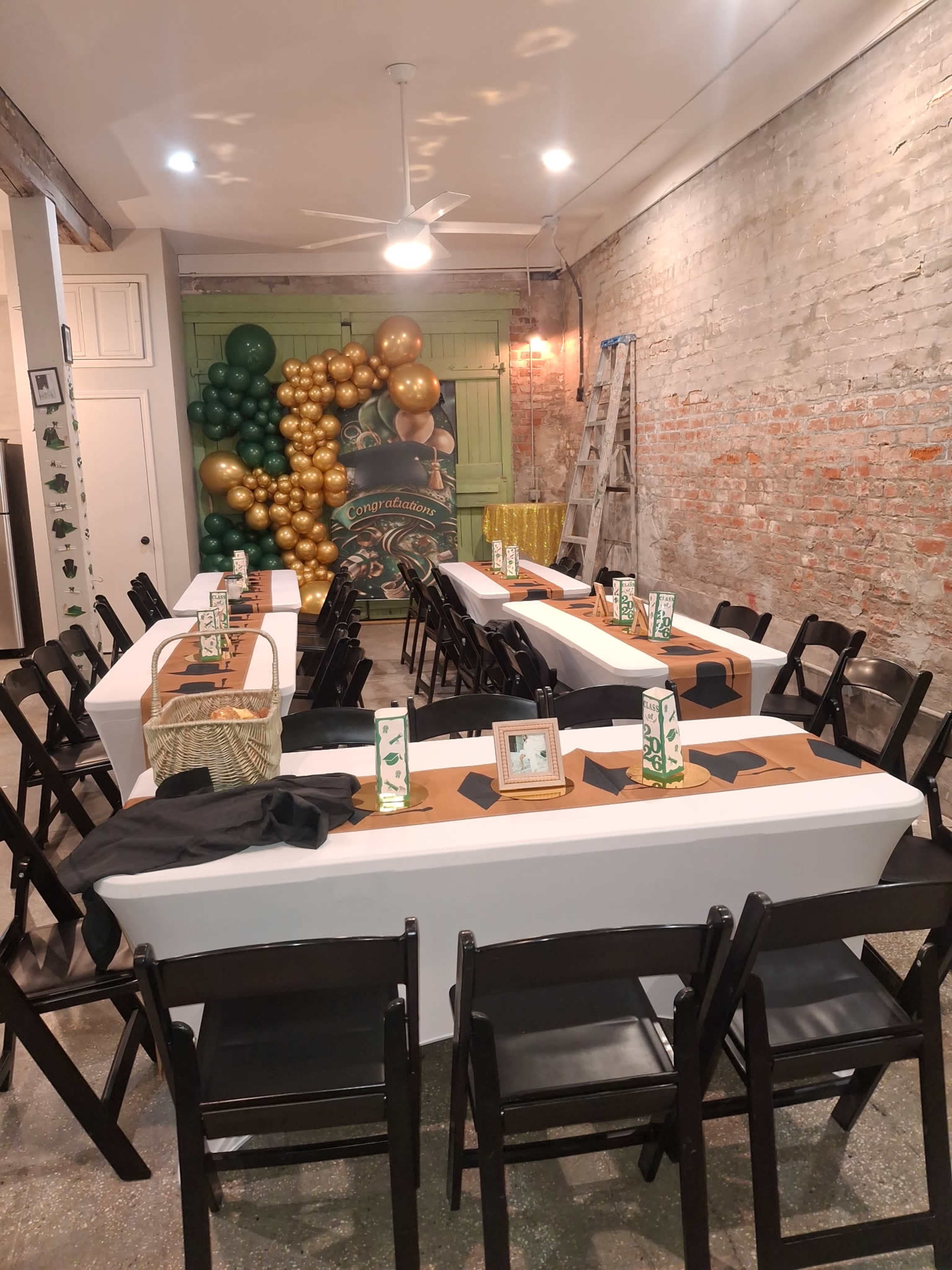 The image depicts a decorated indoor space with multiple tables set up in rows, adorned with brown and green table runners, black napkins, and floral centerpieces, alongside a backdrop of balloons in green and gold.
