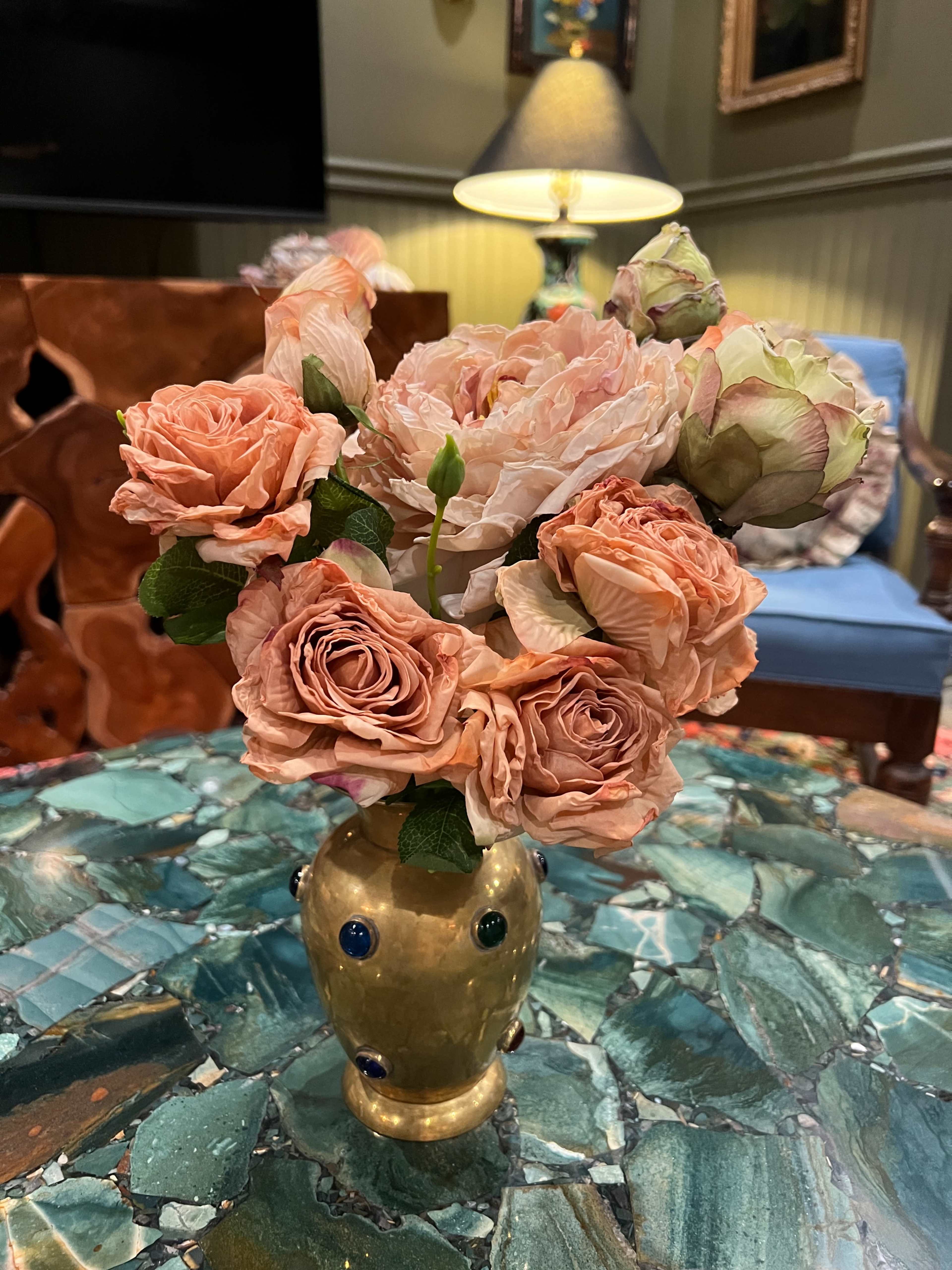 A bouquet of soft-colored roses is arranged in a gold vase on a green stone table, with a blue upholstered chair and lamp in the background.