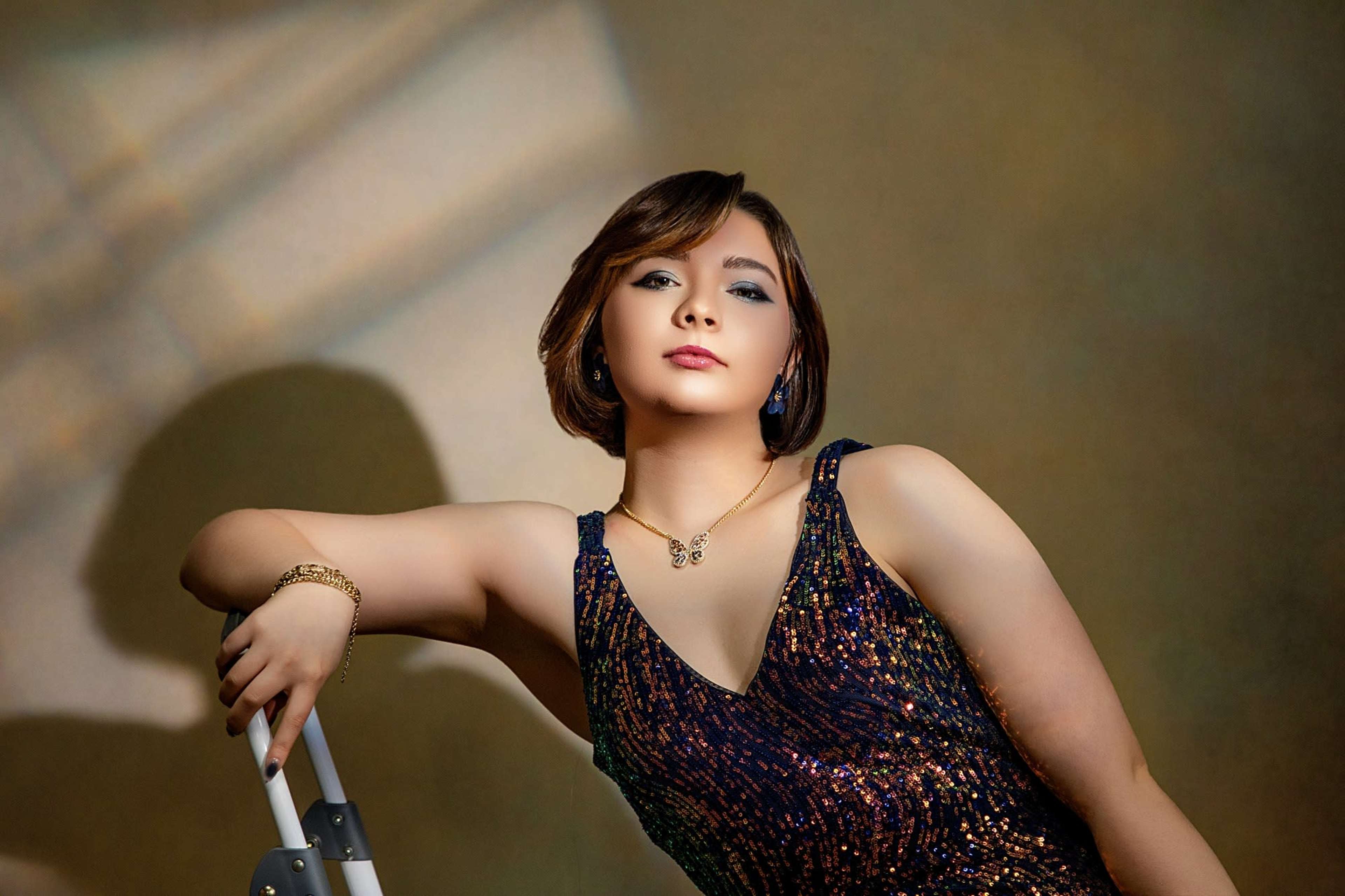 A person wearing a shimmering dress poses confidently with one arm resting on a silver ladder against a softly lit backdrop.