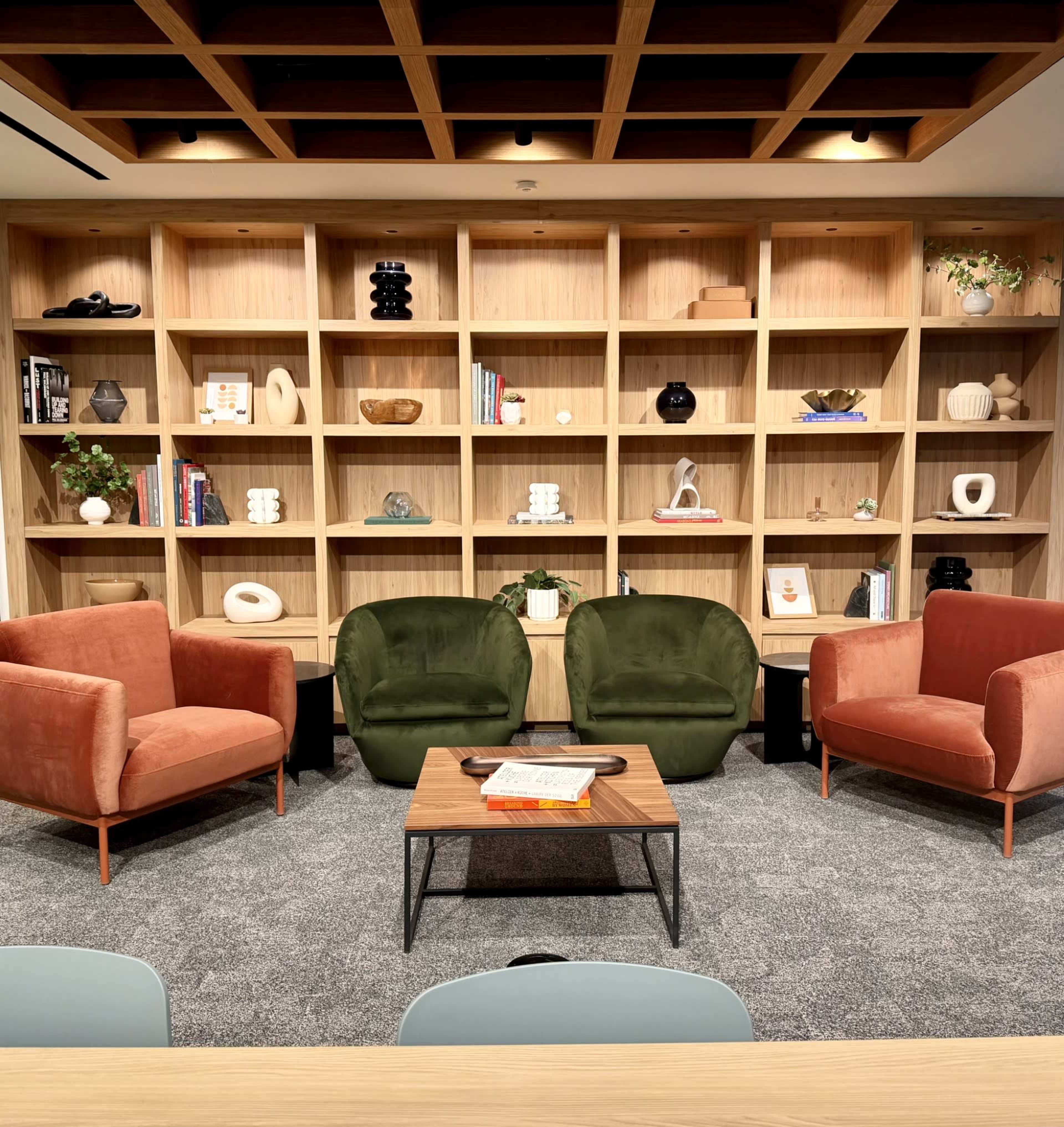 The image shows a cozy lounge area with two upholstered chairs in green and peach, a small wooden coffee table, and a wooden shelving unit filled with various decorative objects and books.