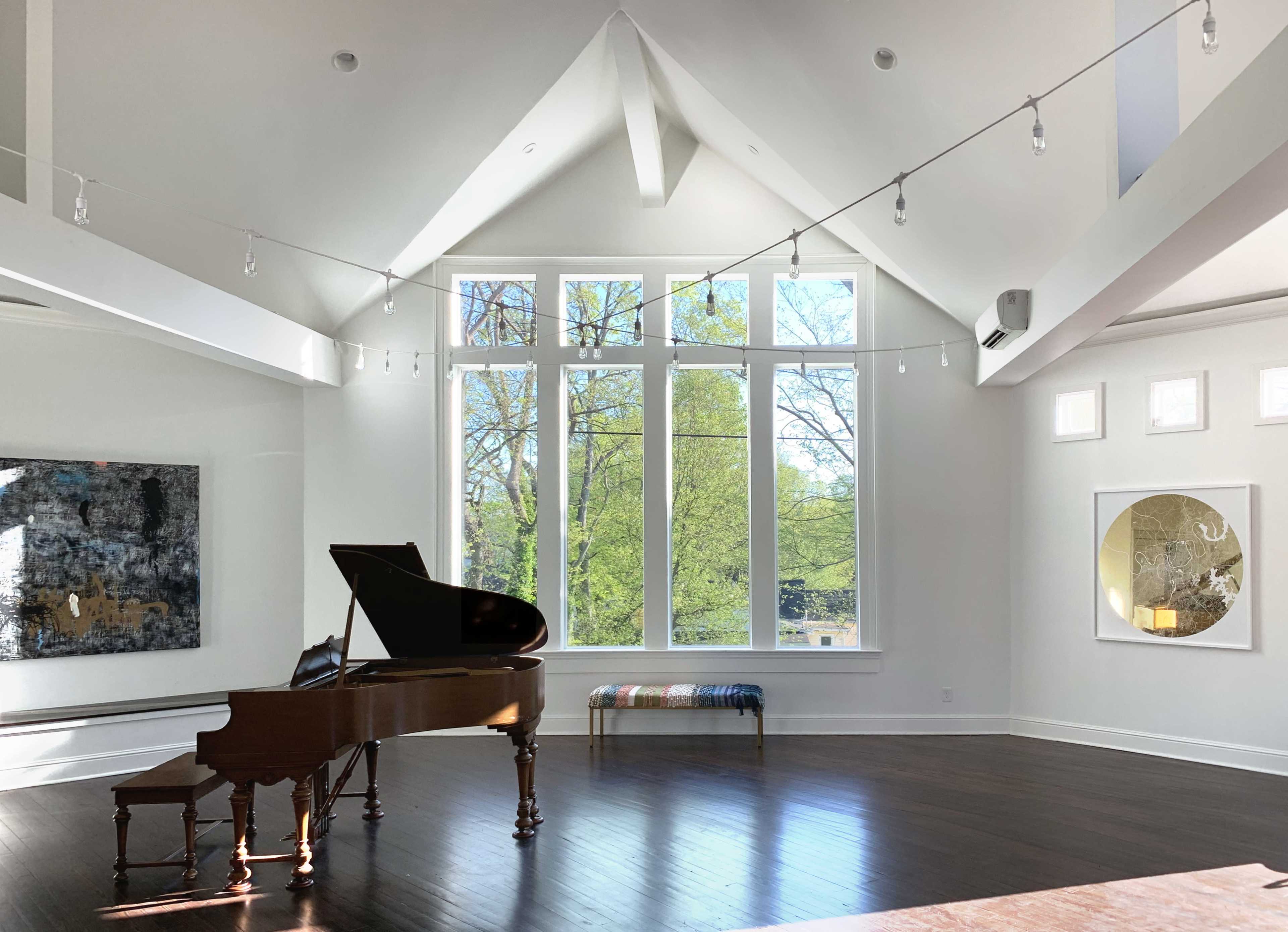 A grand piano is positioned in a brightly lit room featuring large windows that overlook greenery, with contemporary artwork on the walls and a long bench nearby.