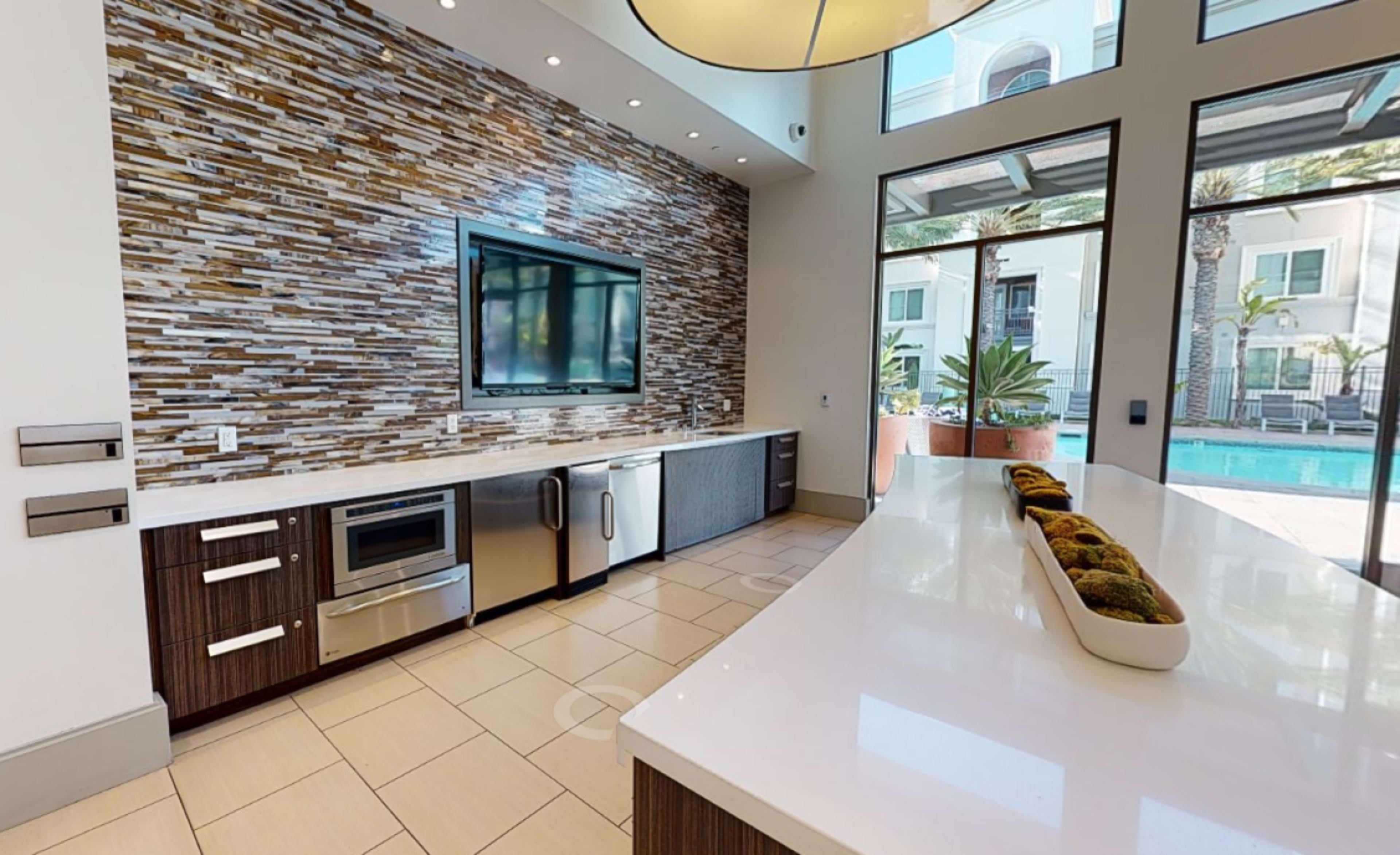 A modern kitchen area with a large countertop, stainless steel appliances, and a stone wall, overlooking a swimming pool through large glass windows.