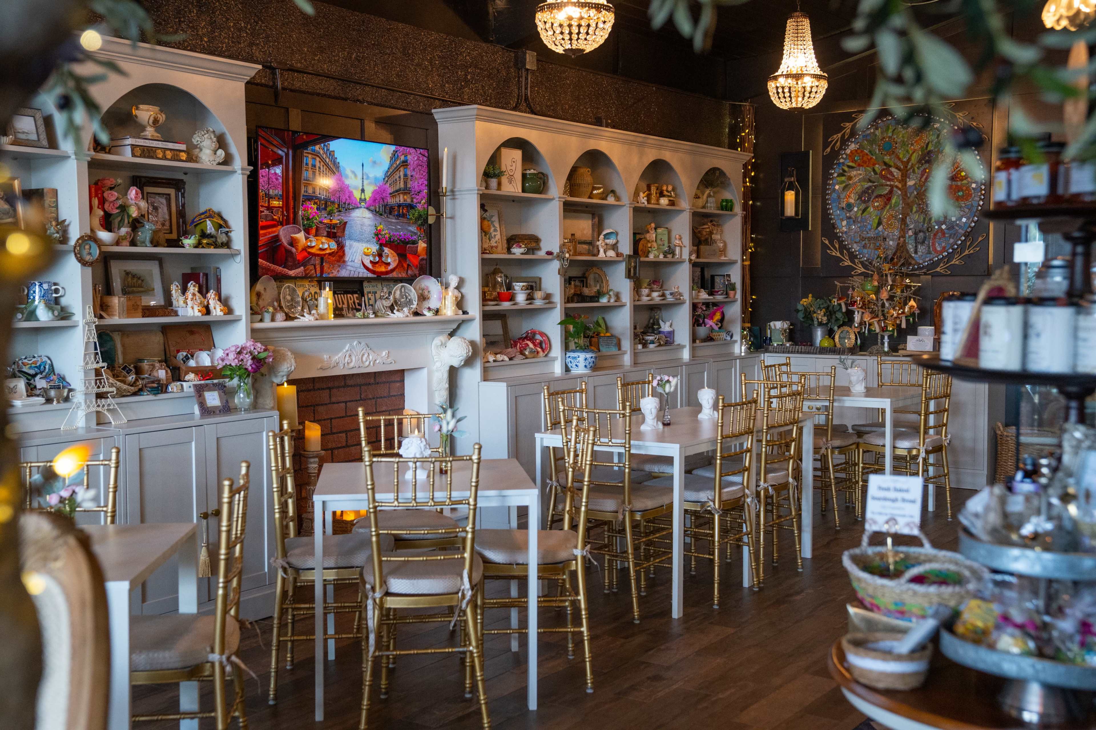 The image shows a charming café with white tables and gold chairs arranged around shelves filled with decorative items and plants.