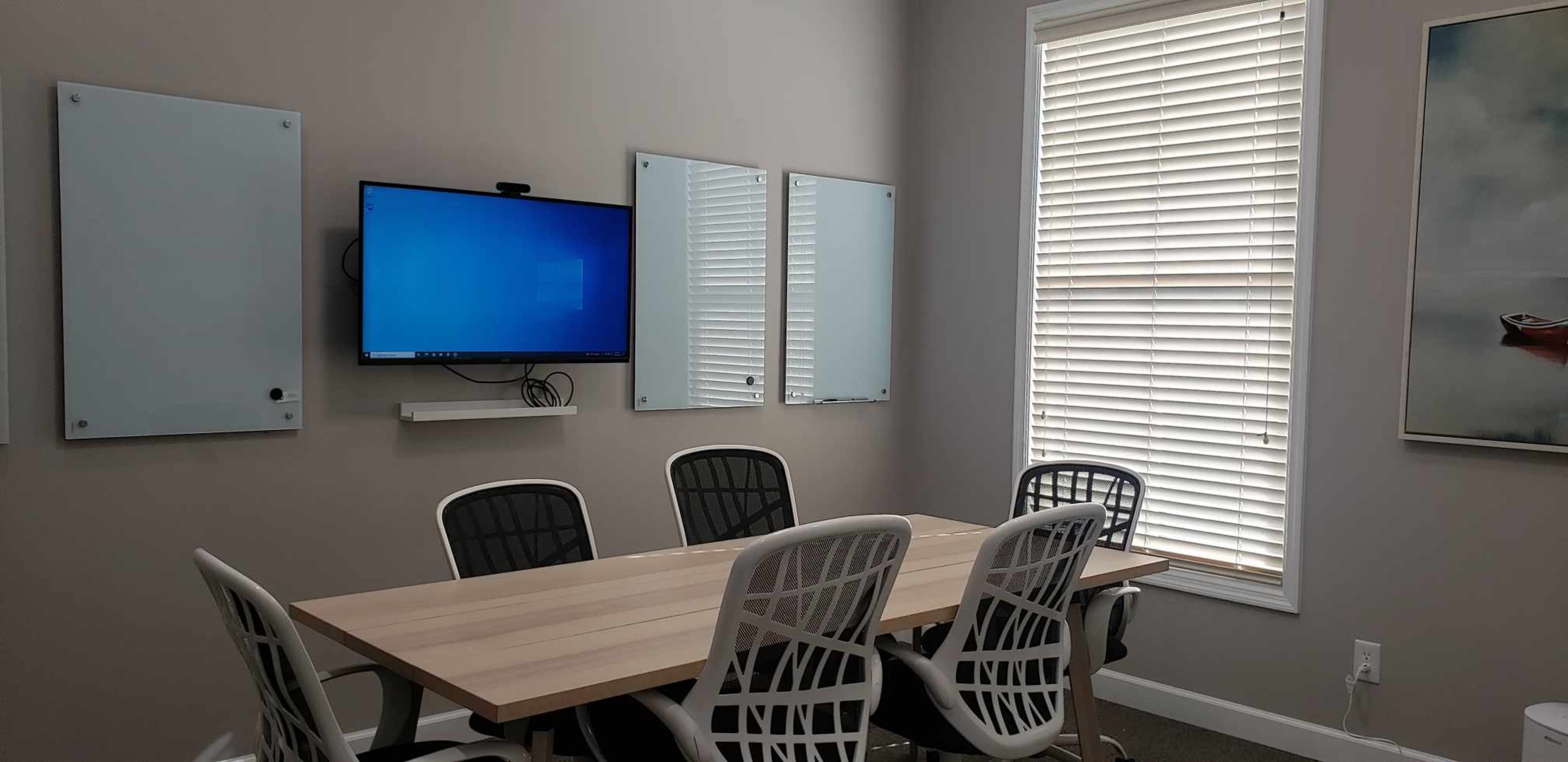 The image shows a modern meeting room featuring a large table surrounded by six white chairs, a television mounted on the wall, and windows with blinds allowing natural light to enter.