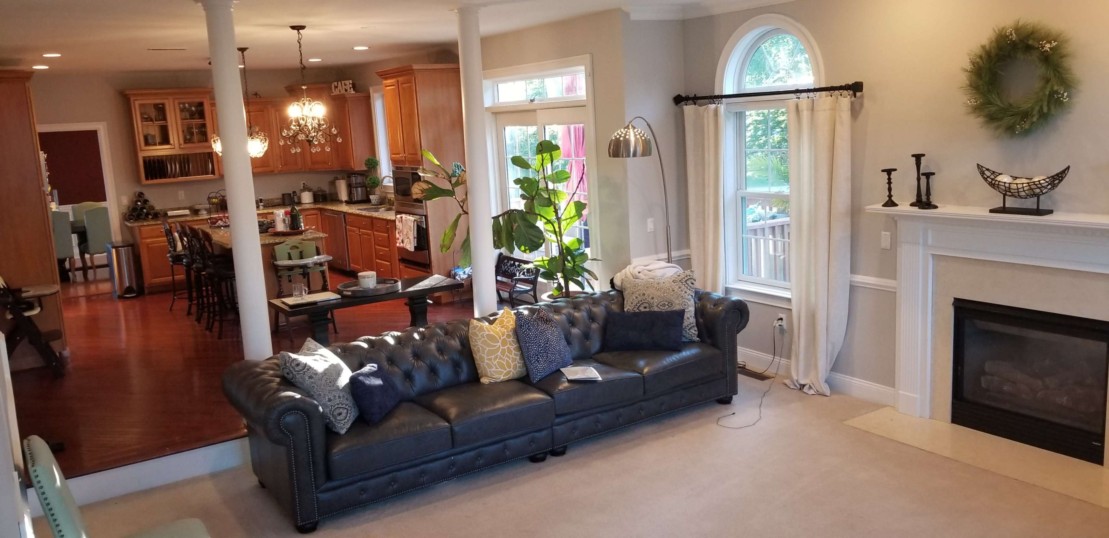The image shows a living room with a leather couch facing a fireplace, while a kitchen and dining area are visible in the background.