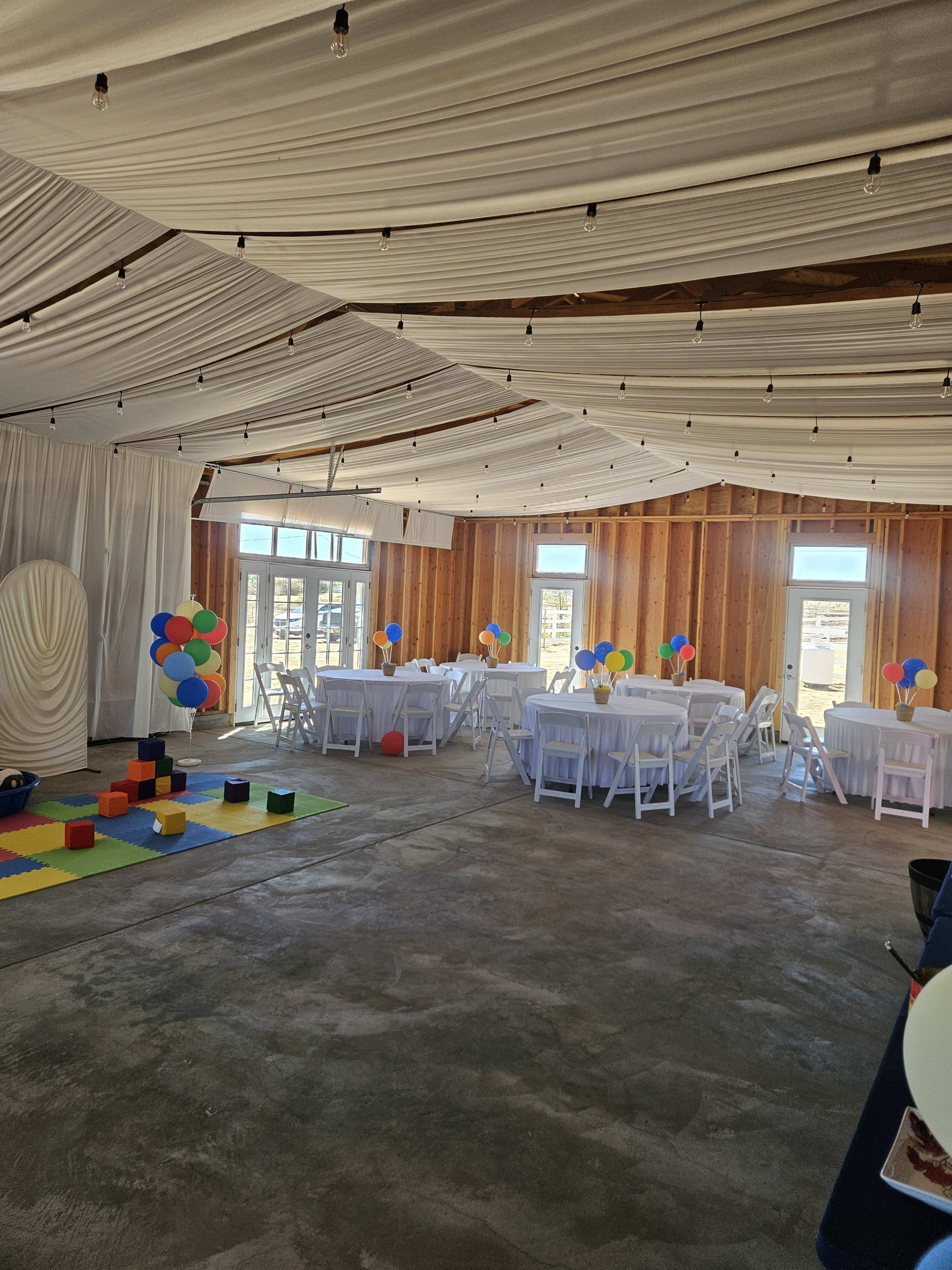 The image shows a spacious event room with white tables and chairs, colorful balloon decorations, and a play area featuring interlocking foam mats and toys.