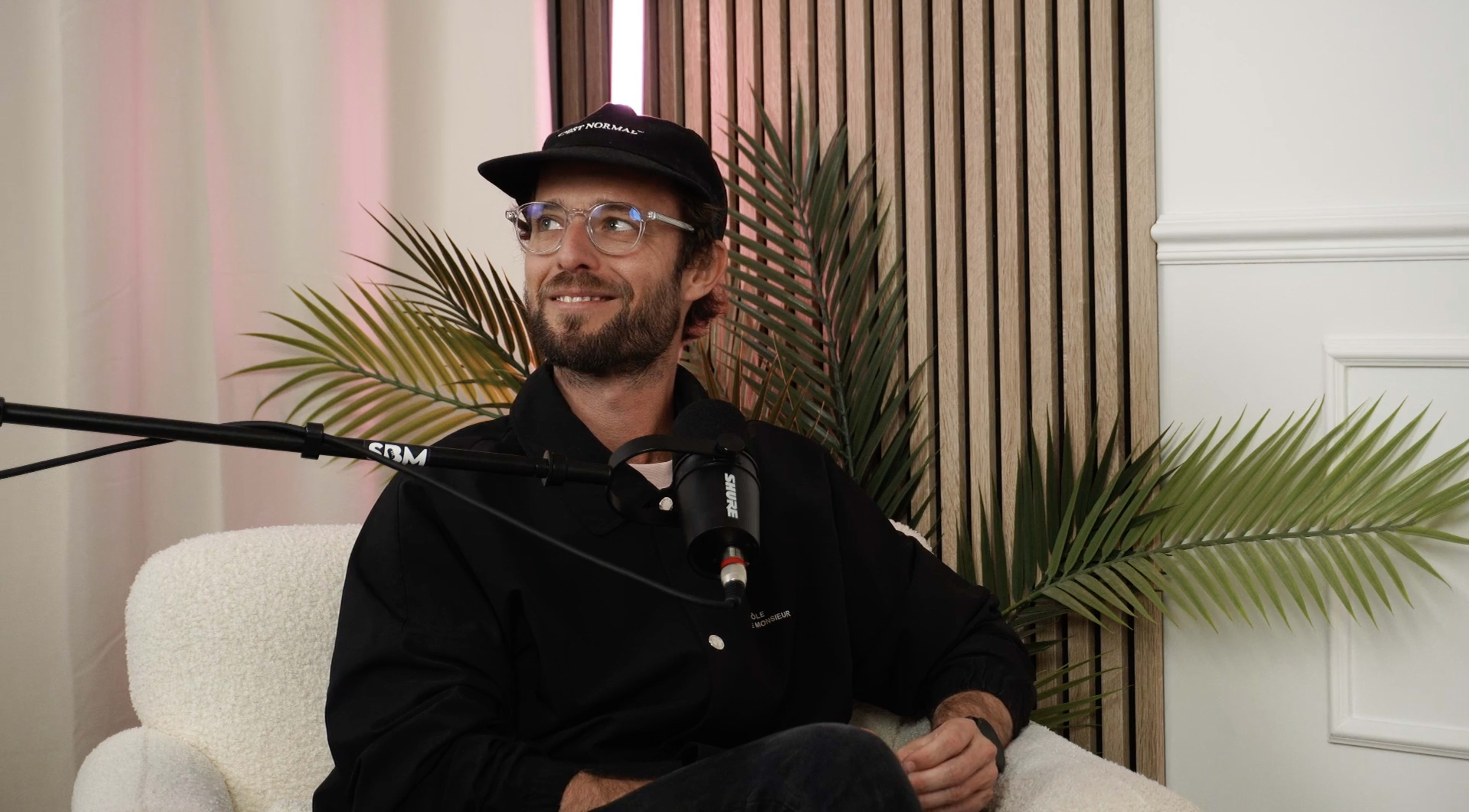 A man wearing a black cap and glasses sits on a light-colored sofa in front of a microphone, surrounded by indoor plants and wooden wall slats.