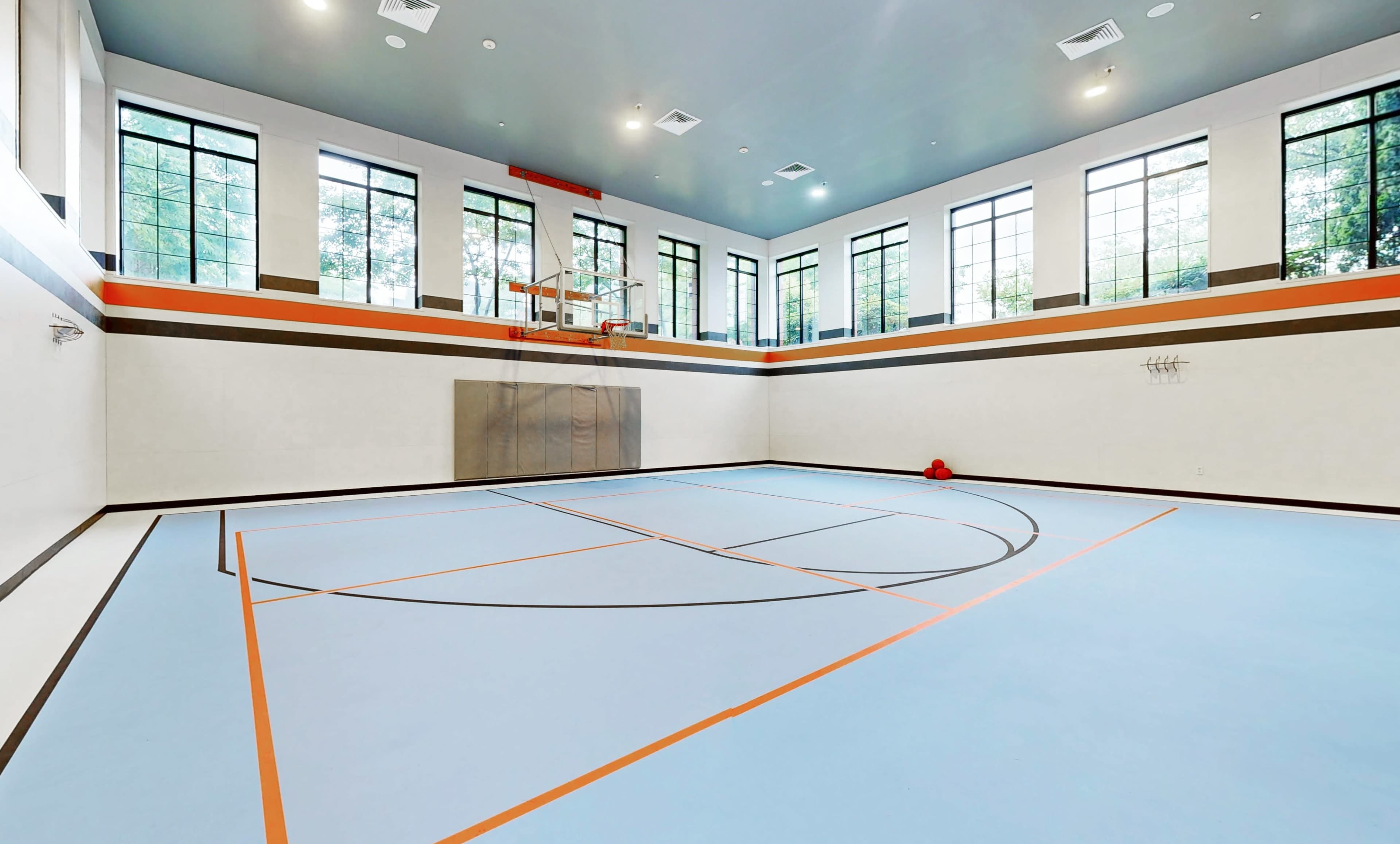 The image shows an empty indoor basketball court with large windows, a hoop at one end, and colored lines marking the playing area on a blue floor.