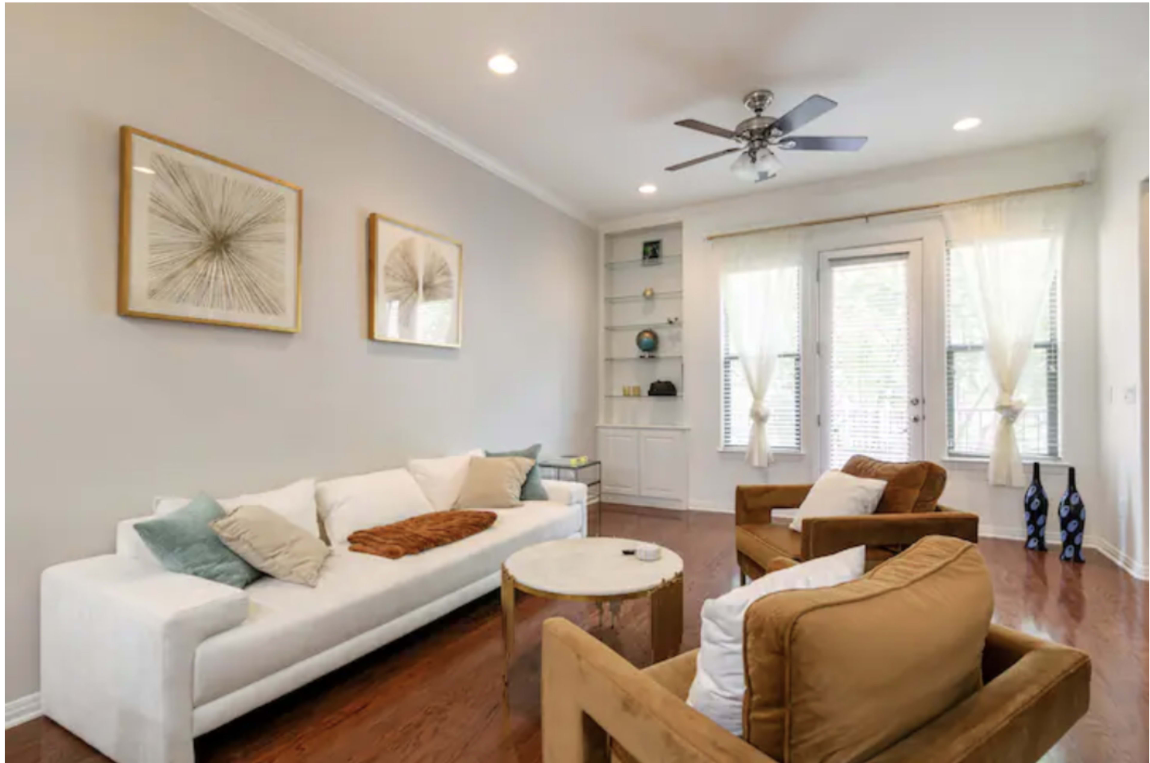 The living room features a white sofa, two brown armchairs, a round wooden coffee table, and framed art on the walls, with large windows allowing natural light.
