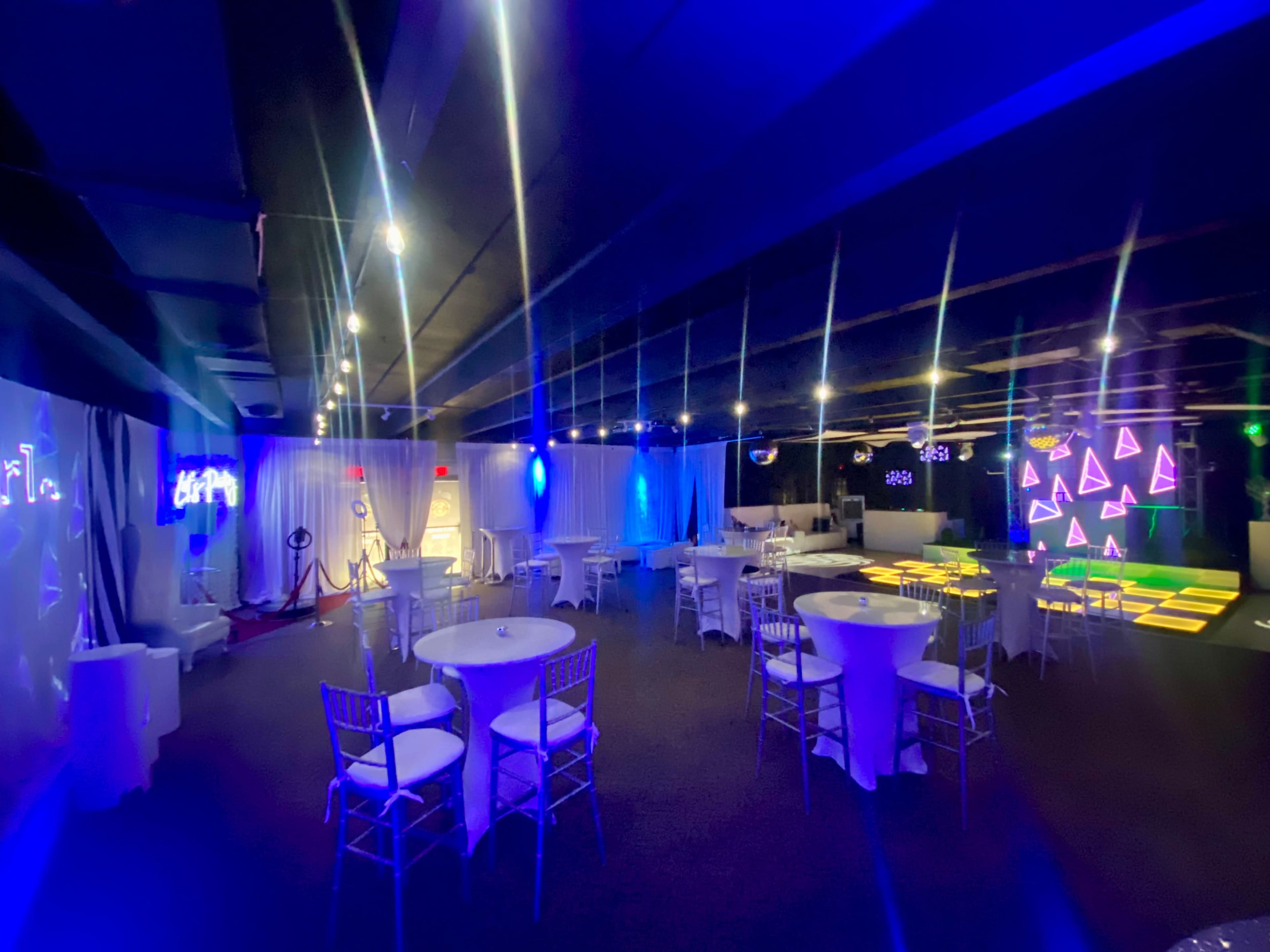 The image shows a dimly lit event space with white tables, chairs, and colorful lighting creating a vibrant atmosphere.
