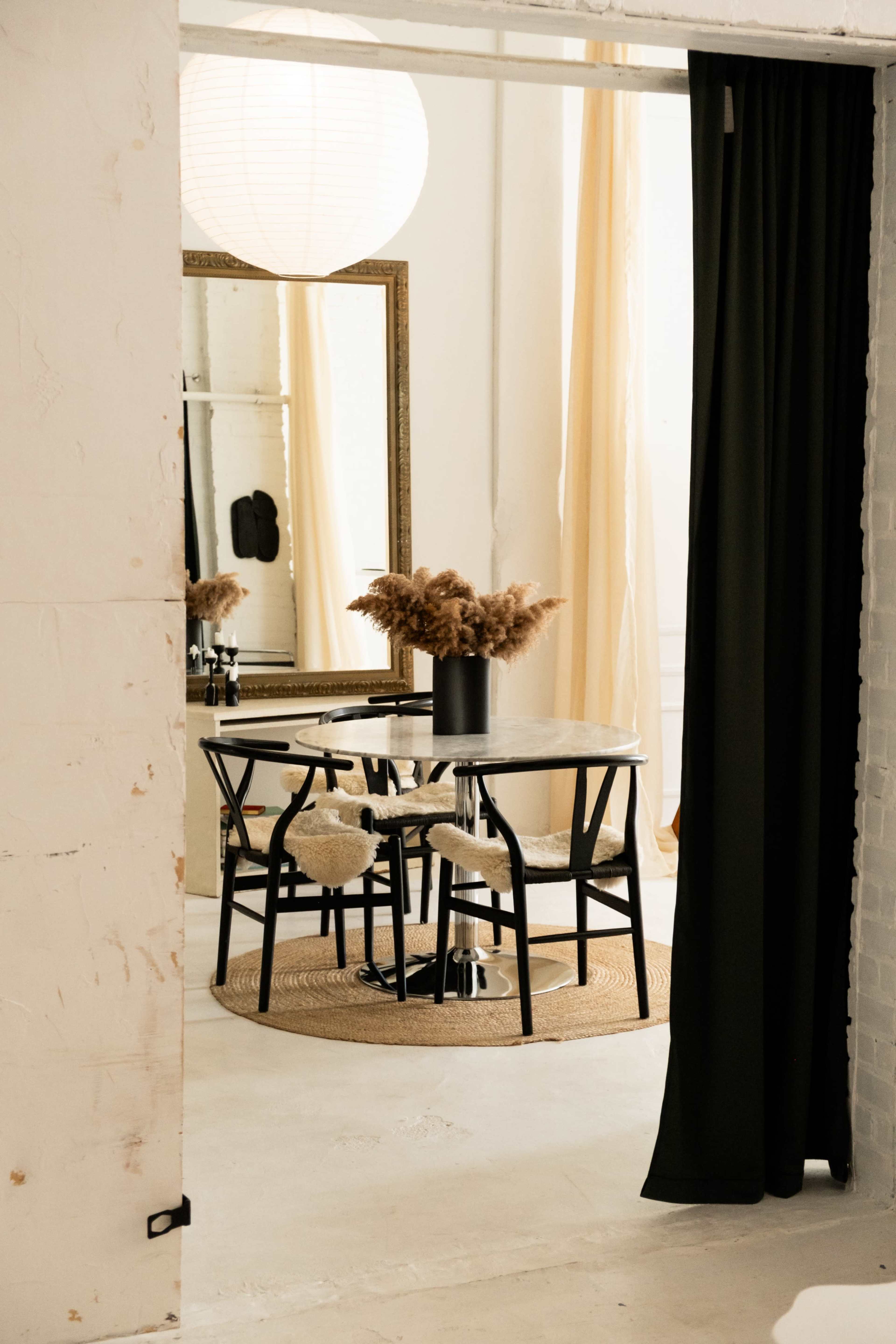 A round dining table with four black chairs is set in a room with a large mirror and soft curtains, complemented by a vase of dried plants.