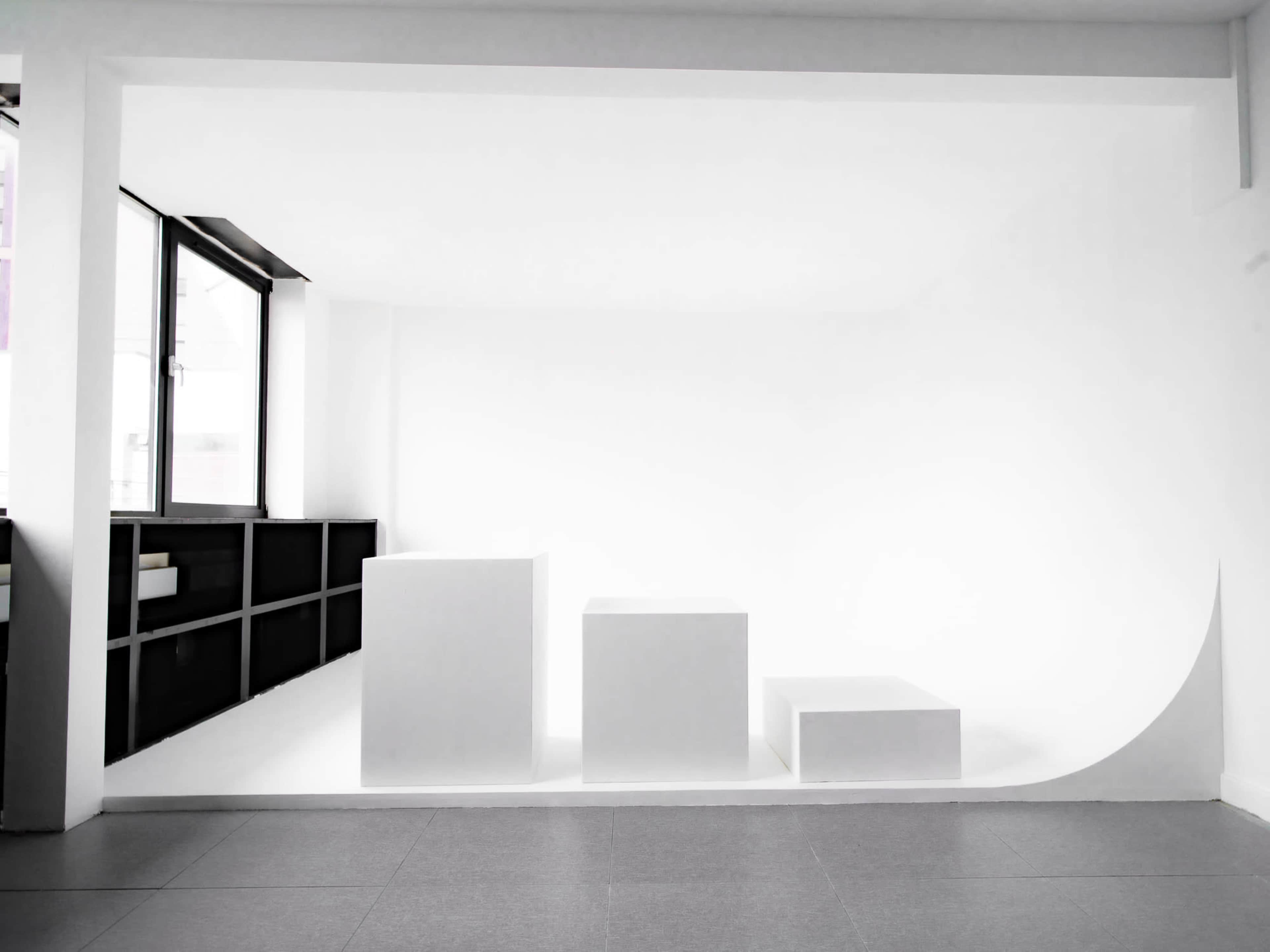 The image shows a minimalist indoor space featuring three white geometric blocks of varying heights against a white backdrop.