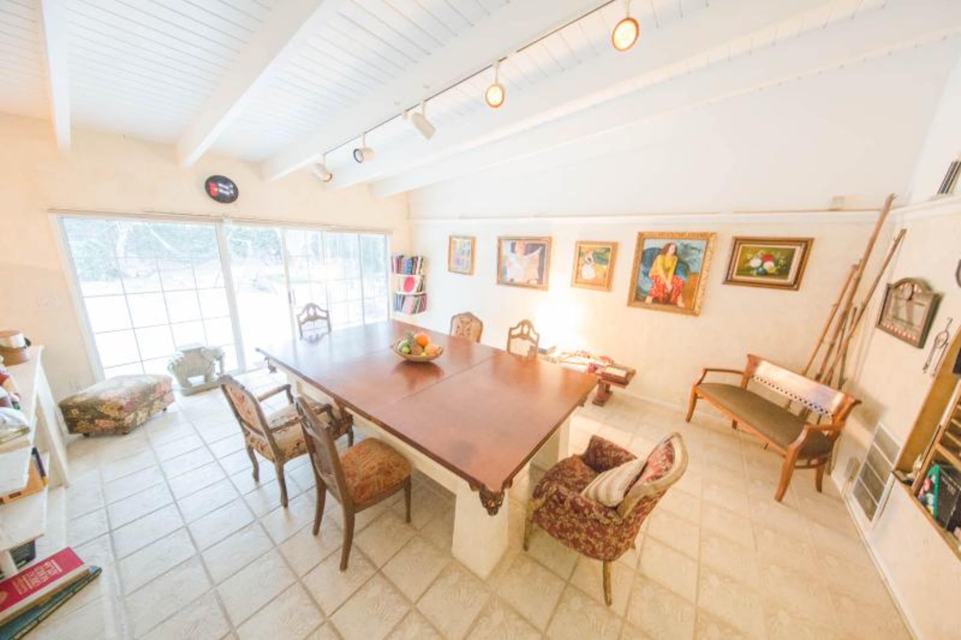 A spacious dining area with a large wooden table surrounded by upholstered chairs, framed artwork on the walls, and large windows overlooking an outdoor space.