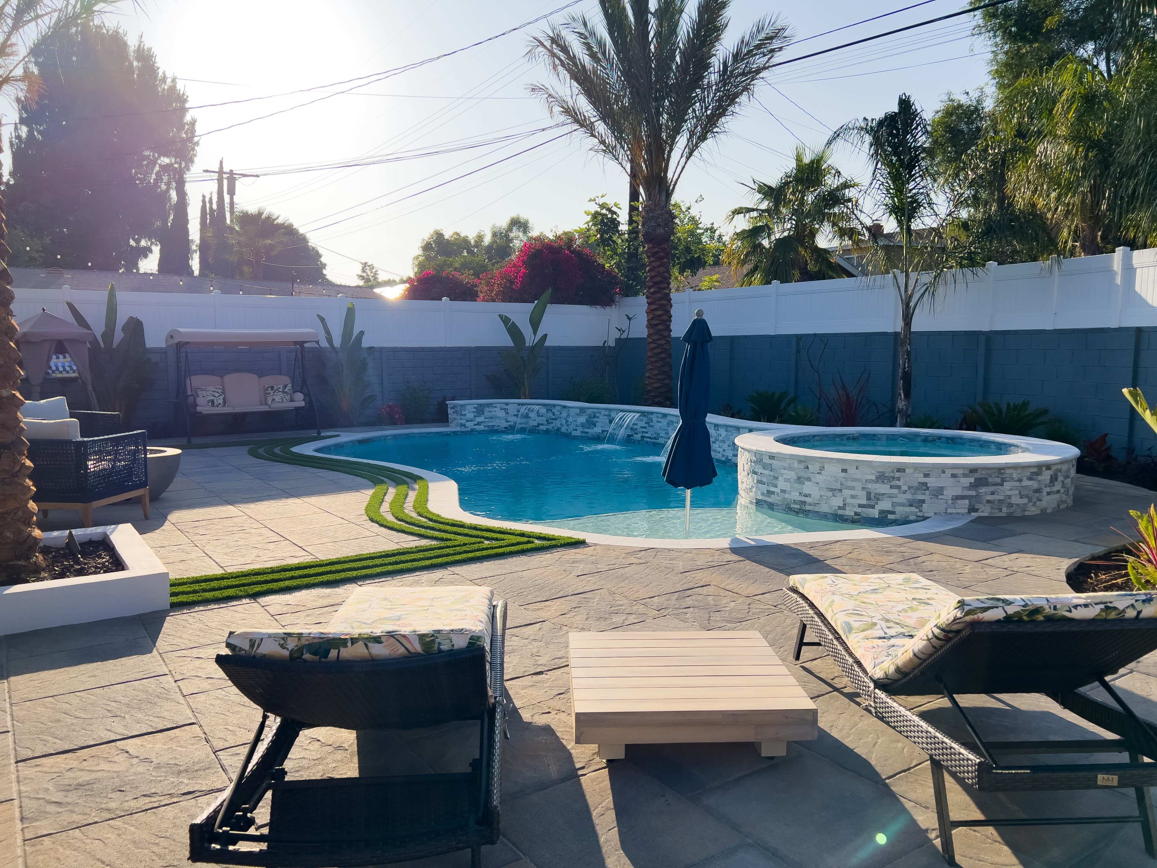A backyard with a swimming pool, a hot tub, two lounge chairs, and tropical landscaping under a clear sky.