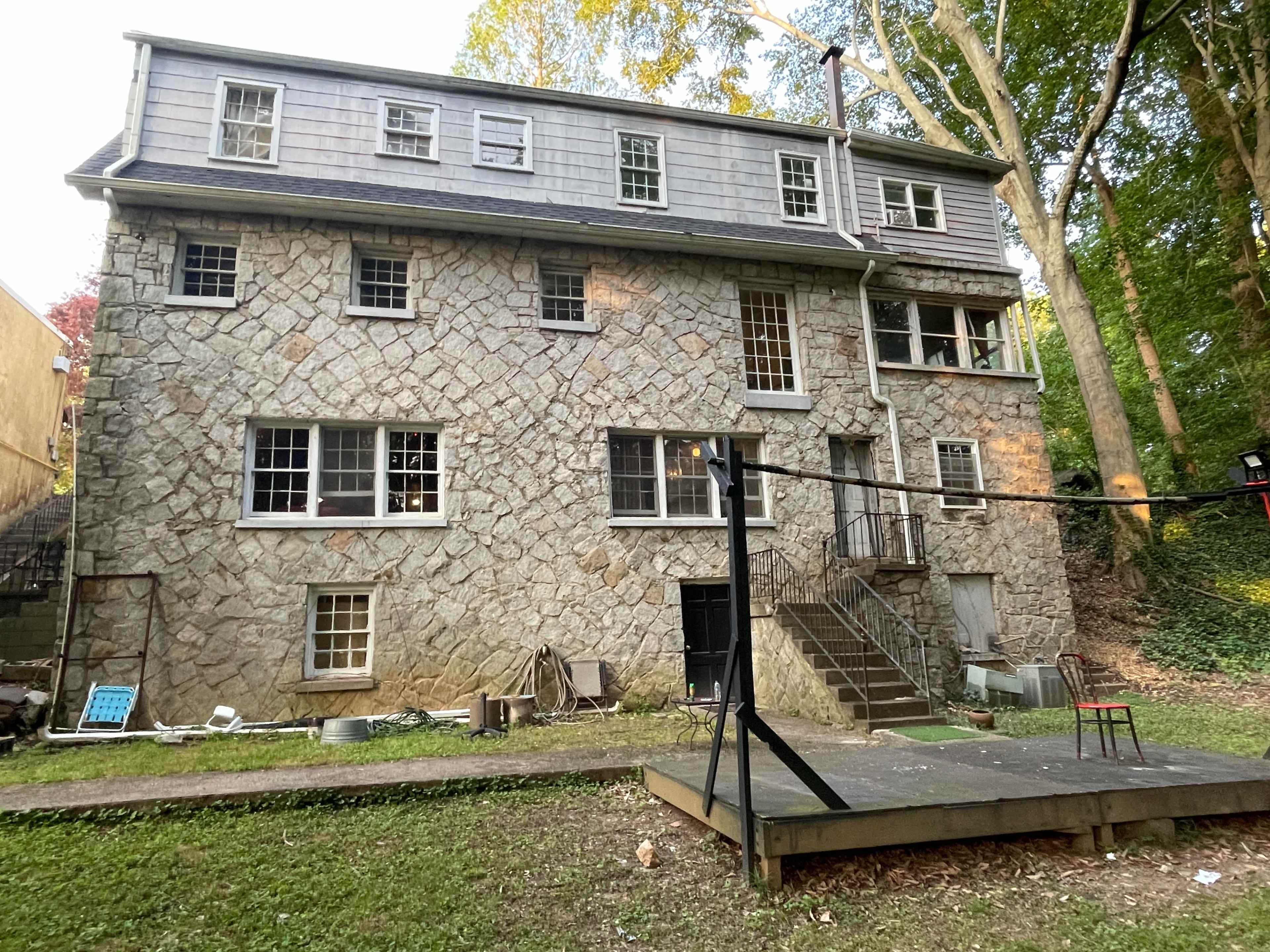 The image shows a three-story stone house with multiple windows and a set of stairs leading to the entrance, surrounded by trees in a wooded area.