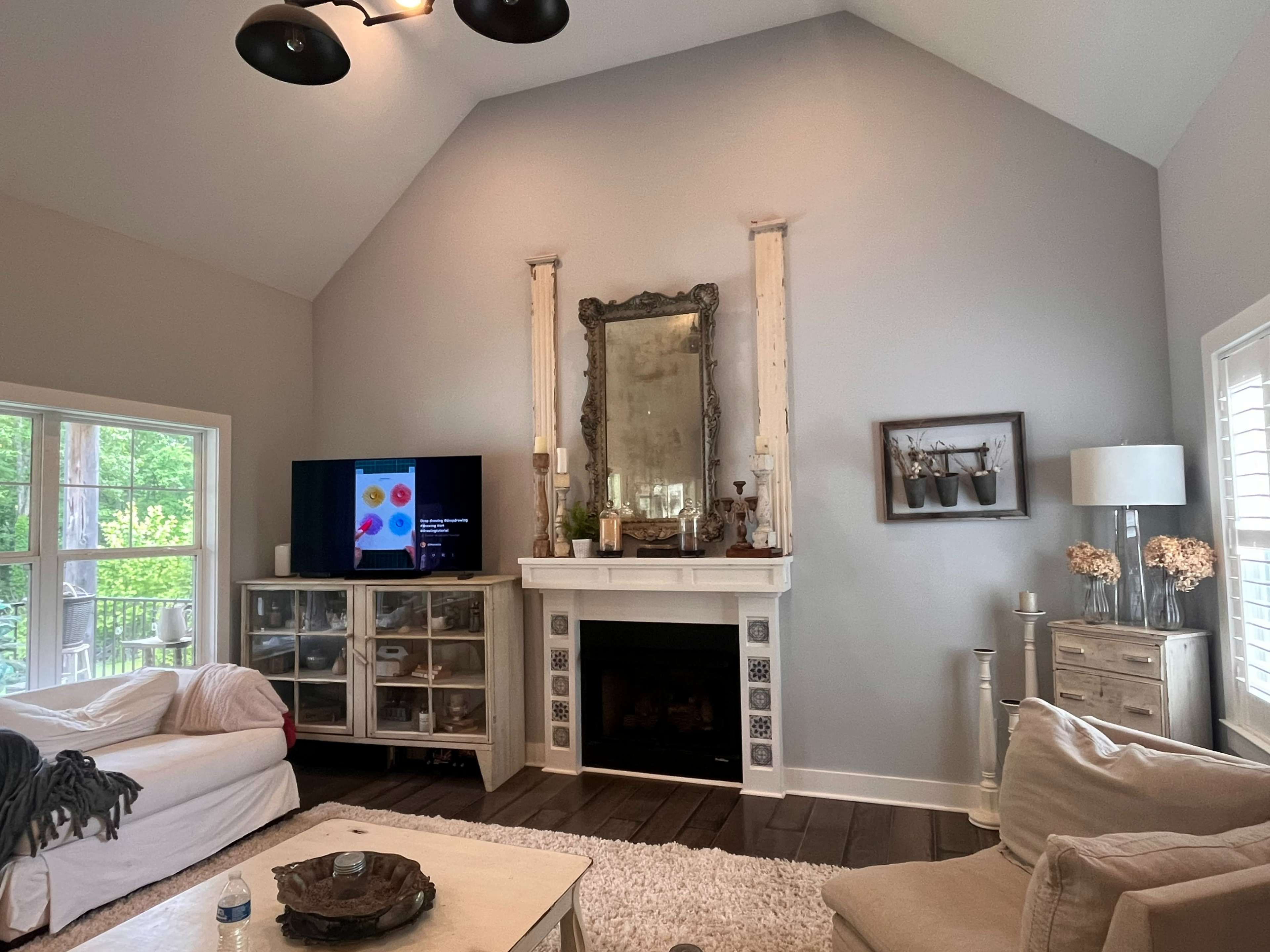 The scene shows a living room with a high ceiling, featuring a fireplace with a large mirror above it, a television on the left, and a light-colored couch opposite a coffee table.