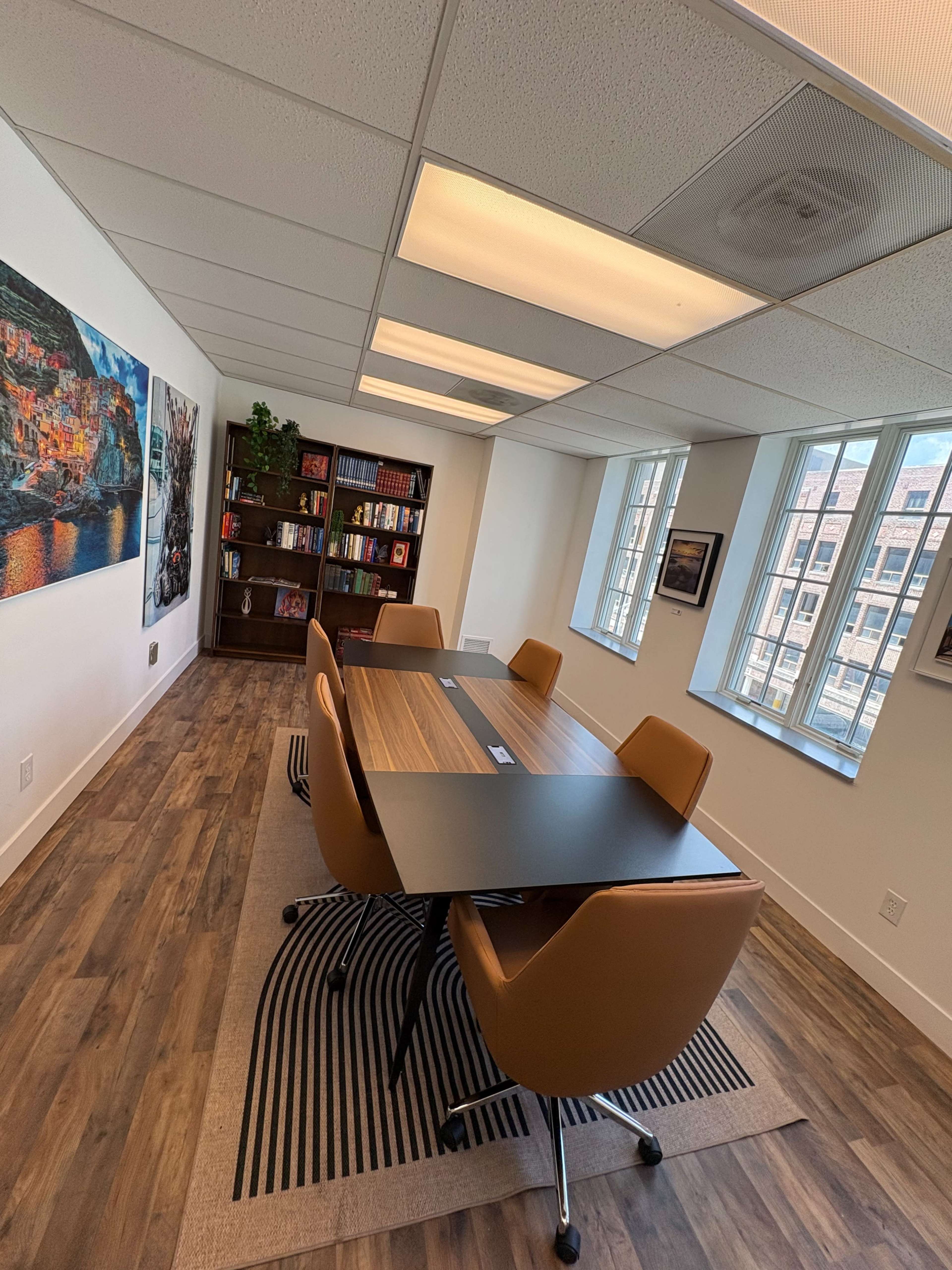 The image shows a bright conference room featuring a long table surrounded by tan chairs, with a bookshelf filled with books and a large wall art piece.