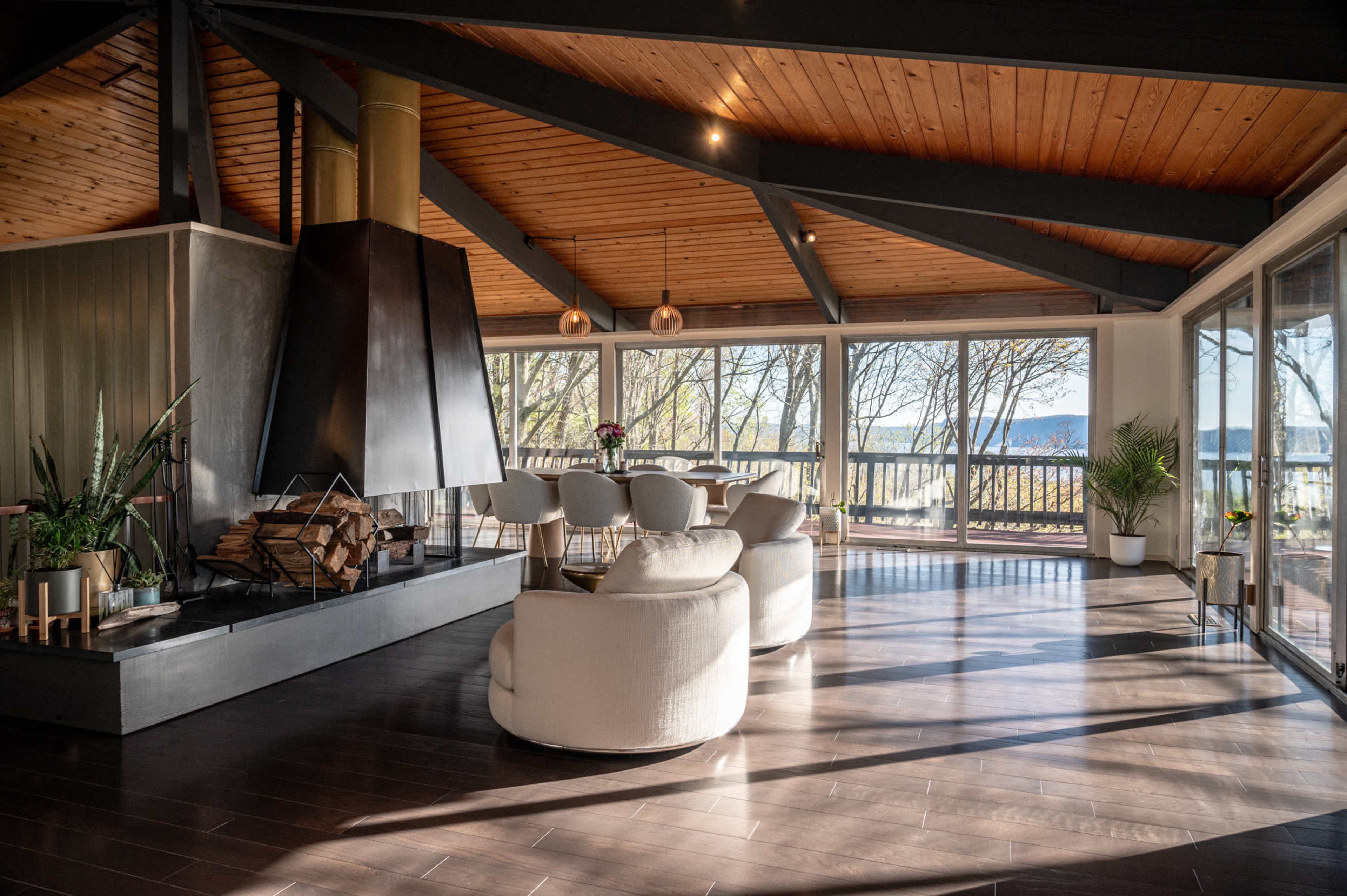 The interior features a modern open-plan living space with a large fireplace, circular sofas, and a dining area with views of a tree-lined outdoor setting.