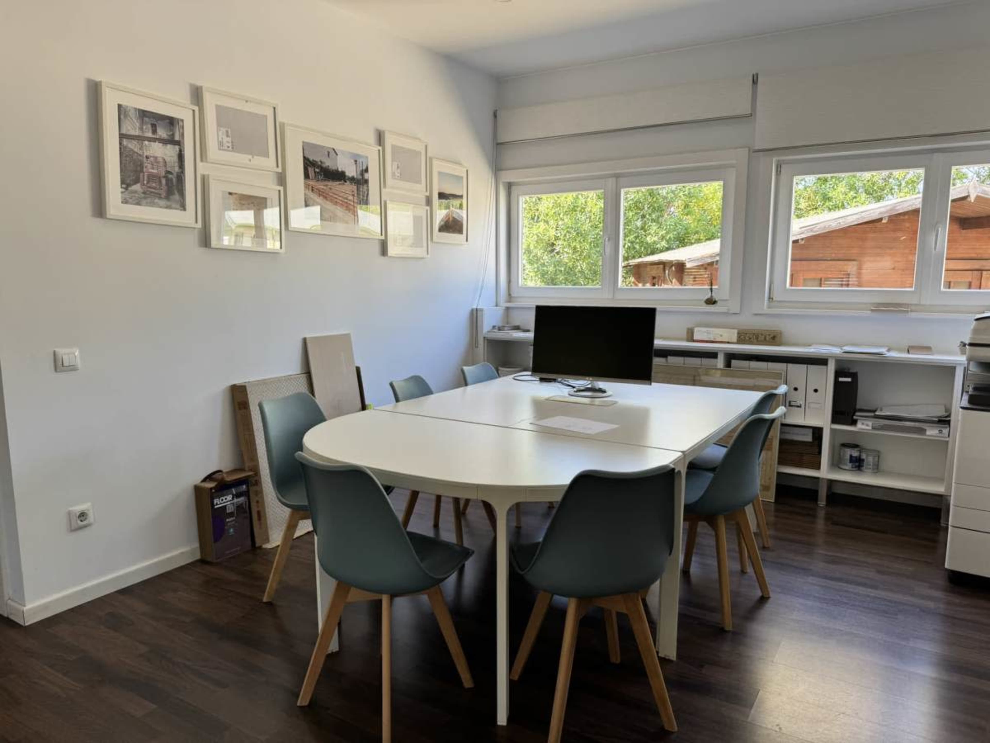 The image shows a modern workspace with a round white table, multiple chairs, and framed photographs on the walls.