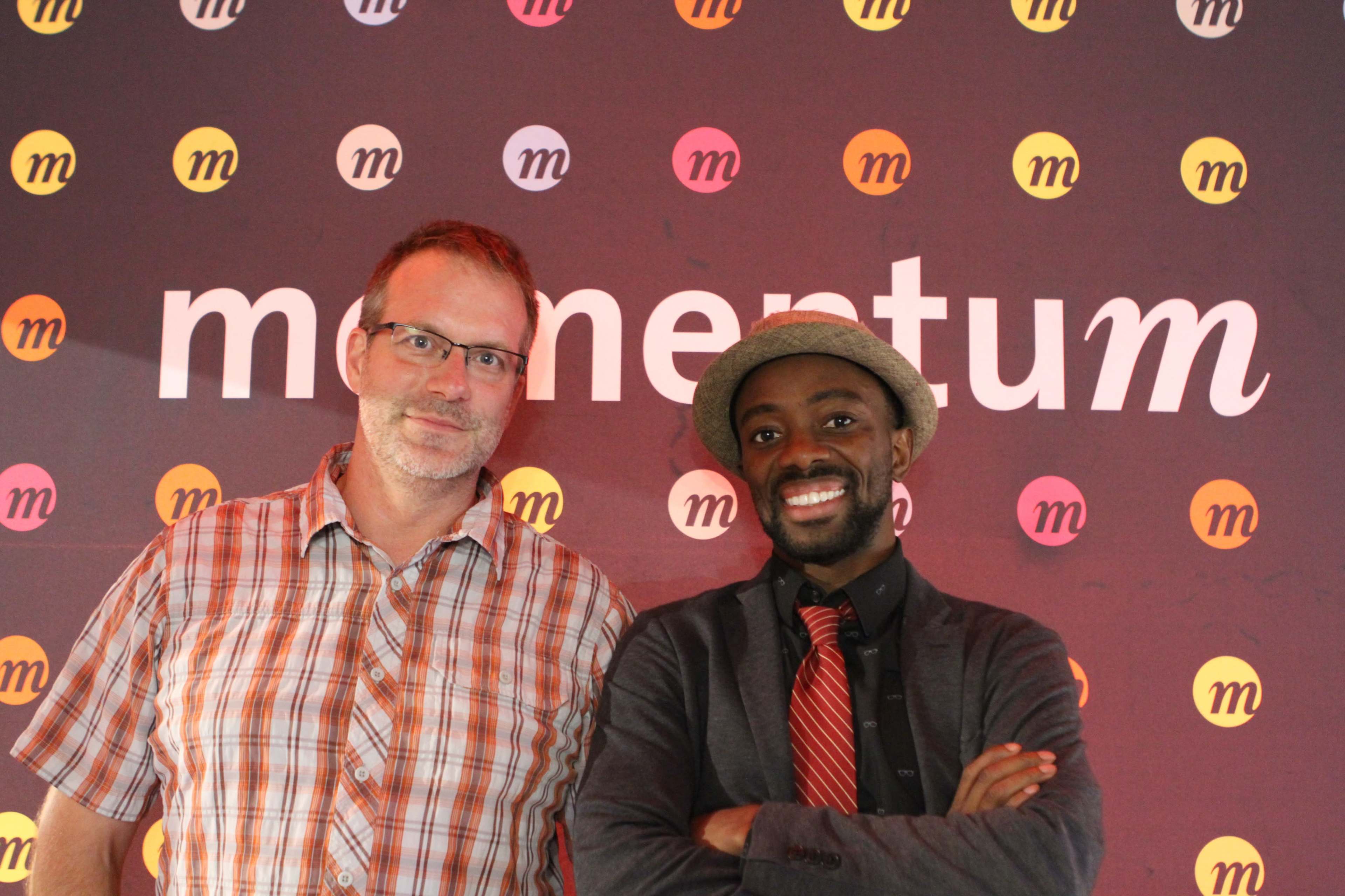 Two men pose for a photo in front of a backdrop displaying the word "momentum" with colorful circular logos.
