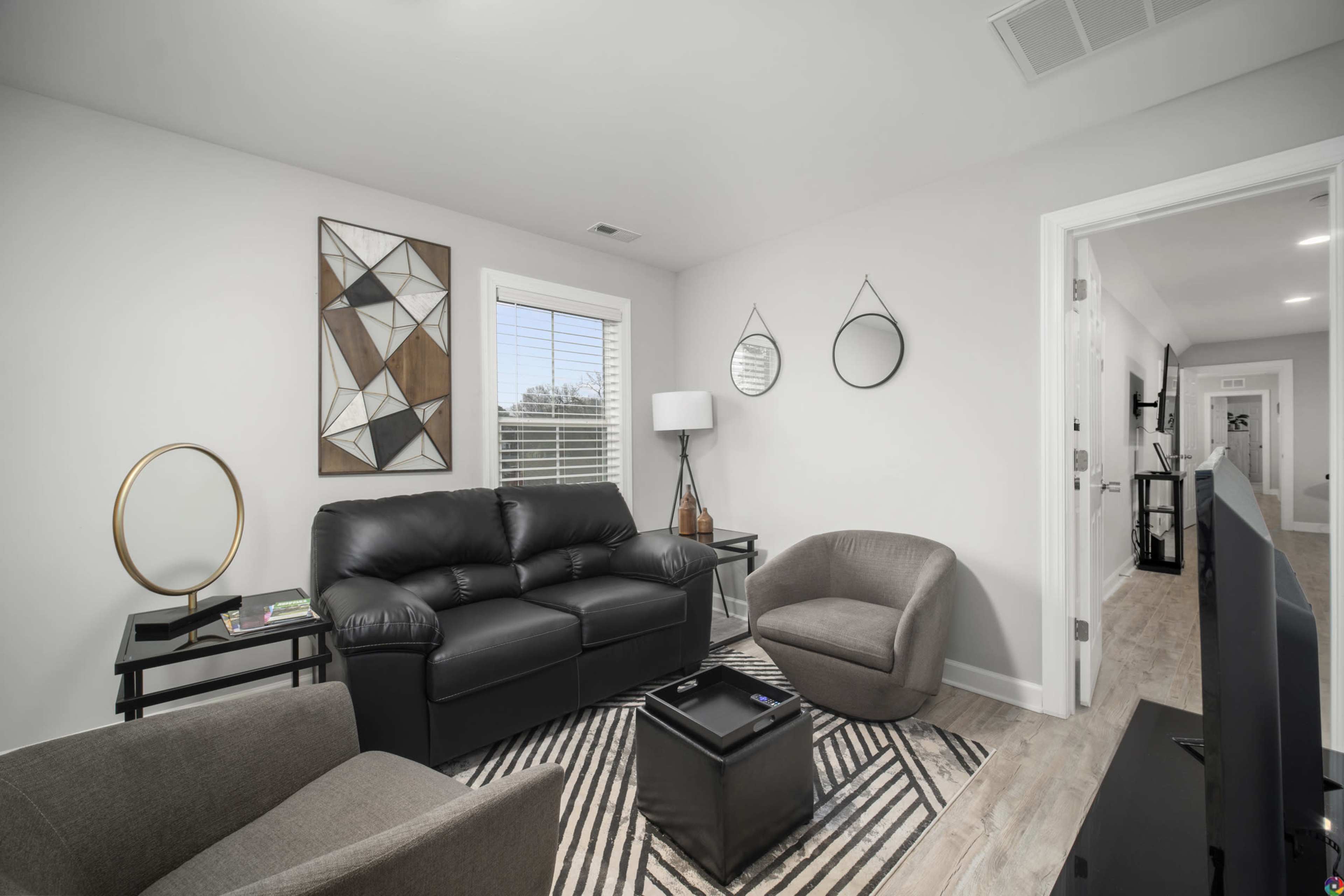 A modern living room featuring a black leather sofa, two upholstered chairs, a geometric wall art piece, and a TV stand against a neutral wall.