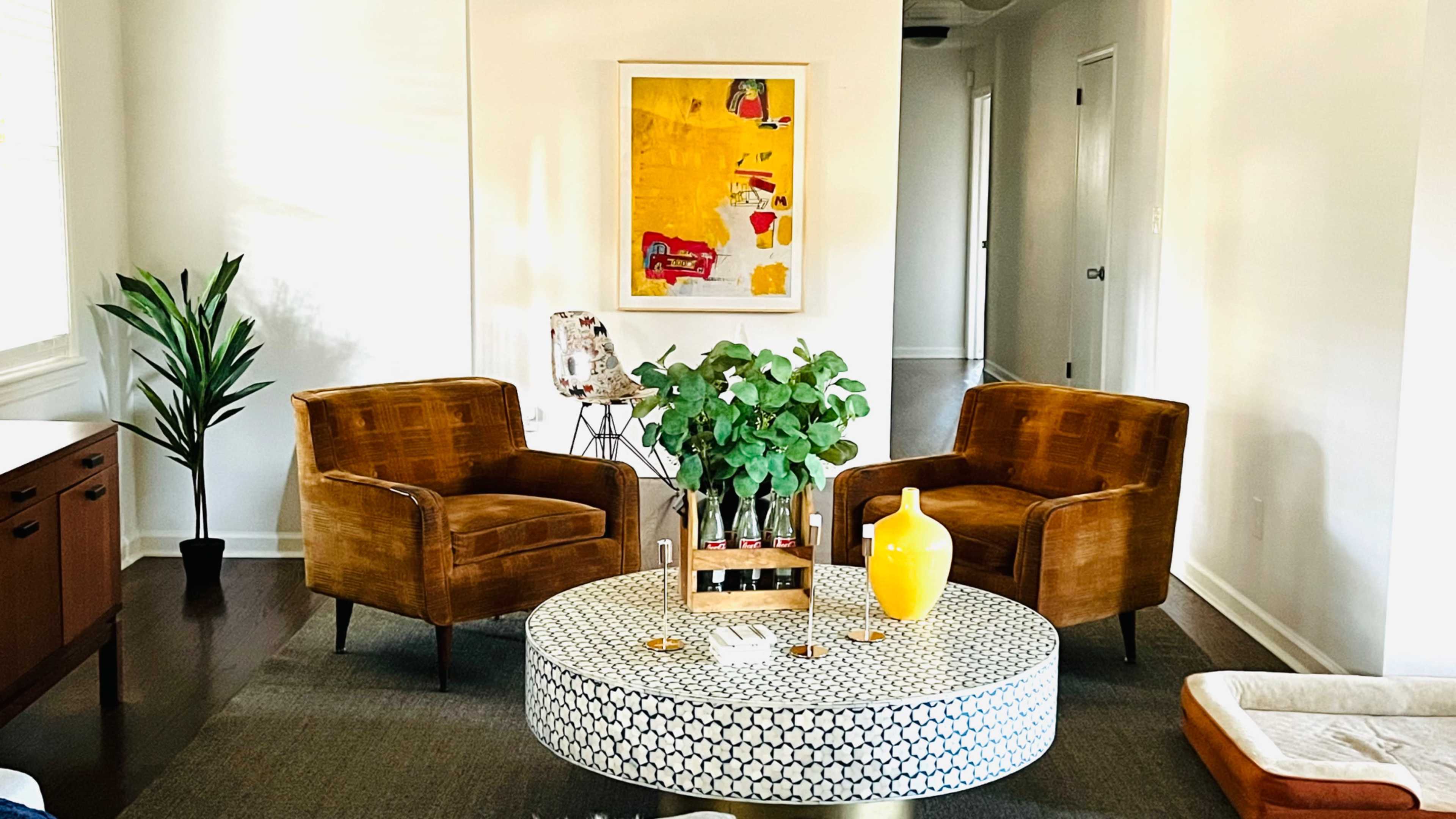 The image shows a modern living room with two brown chairs, a round coffee table with a patterned surface, a potted plant, and a yellow decorative vase.