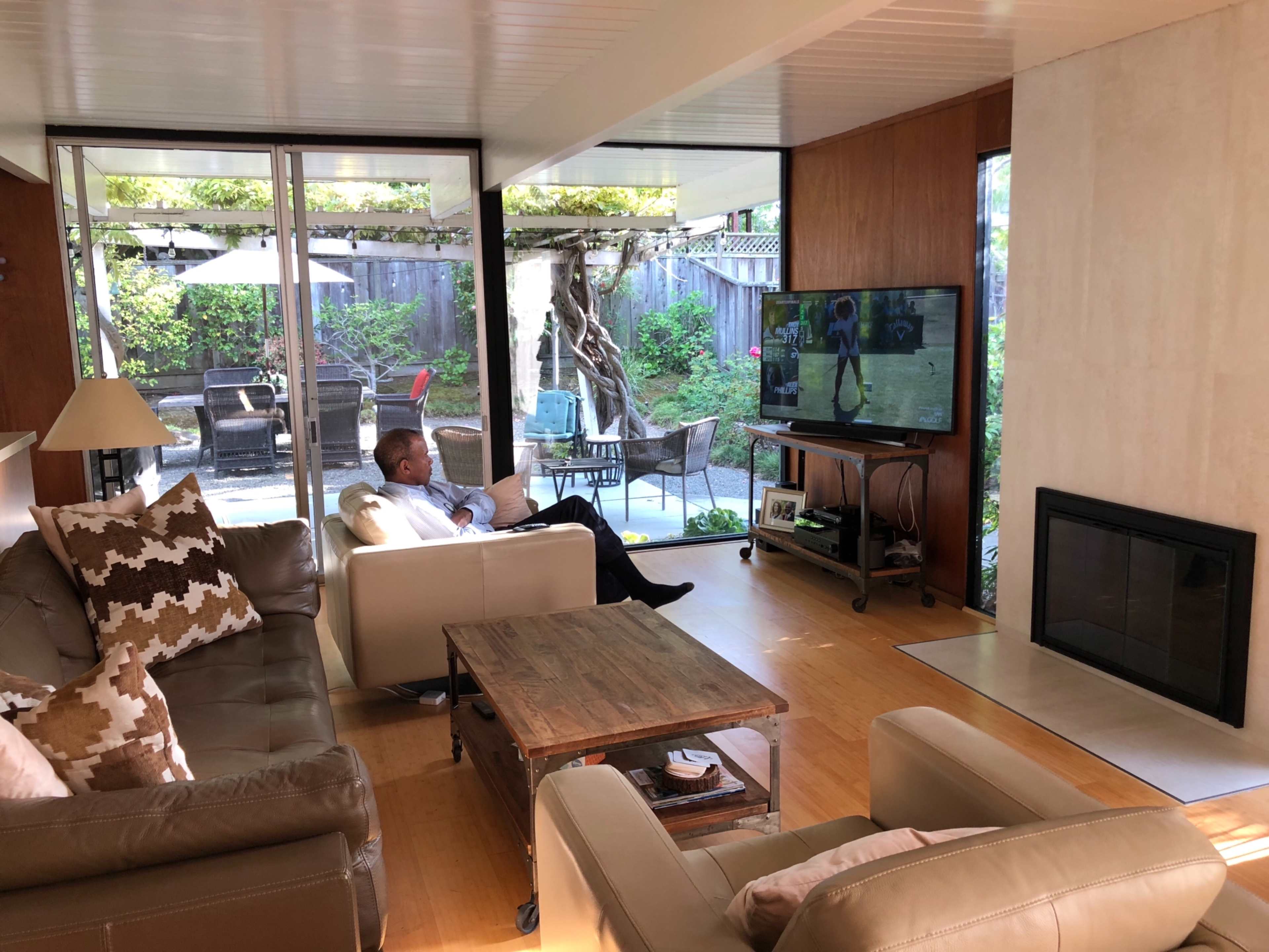 A man sits on a couch watching television in a modern living room with large windows overlooking a patio.