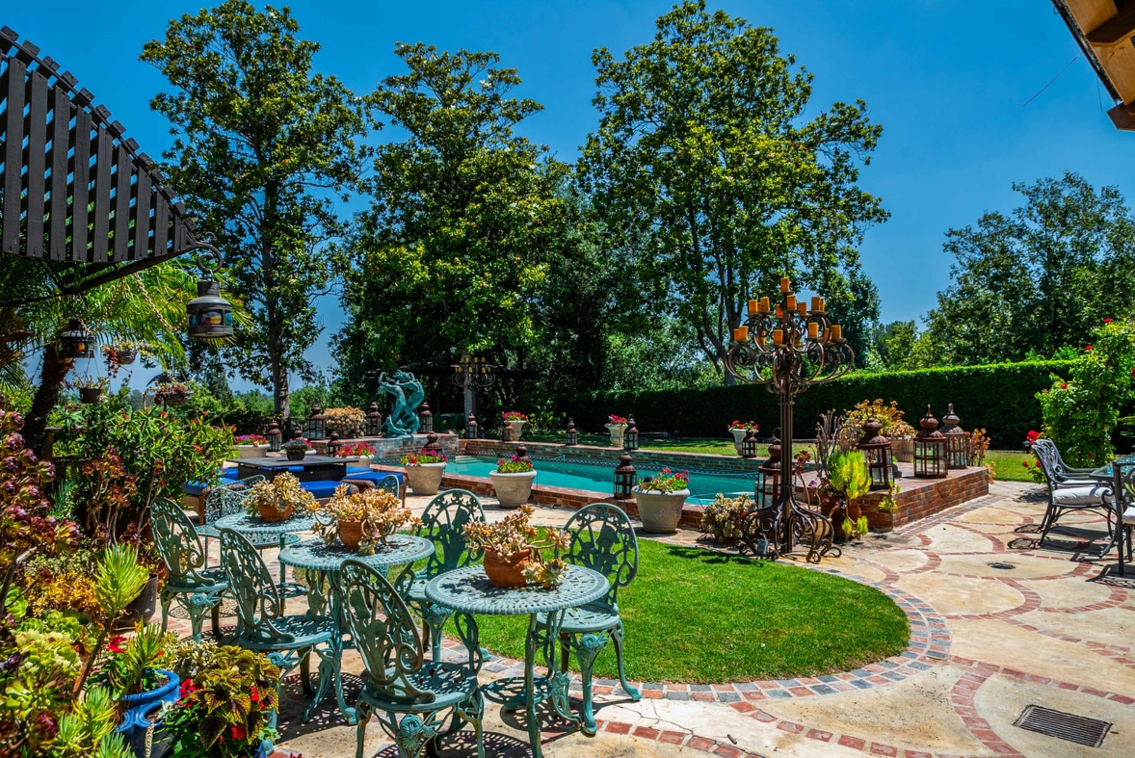 The image shows a garden area with a swimming pool, surrounded by trees and decorated with various plants, sculptures, and outdoor furniture.