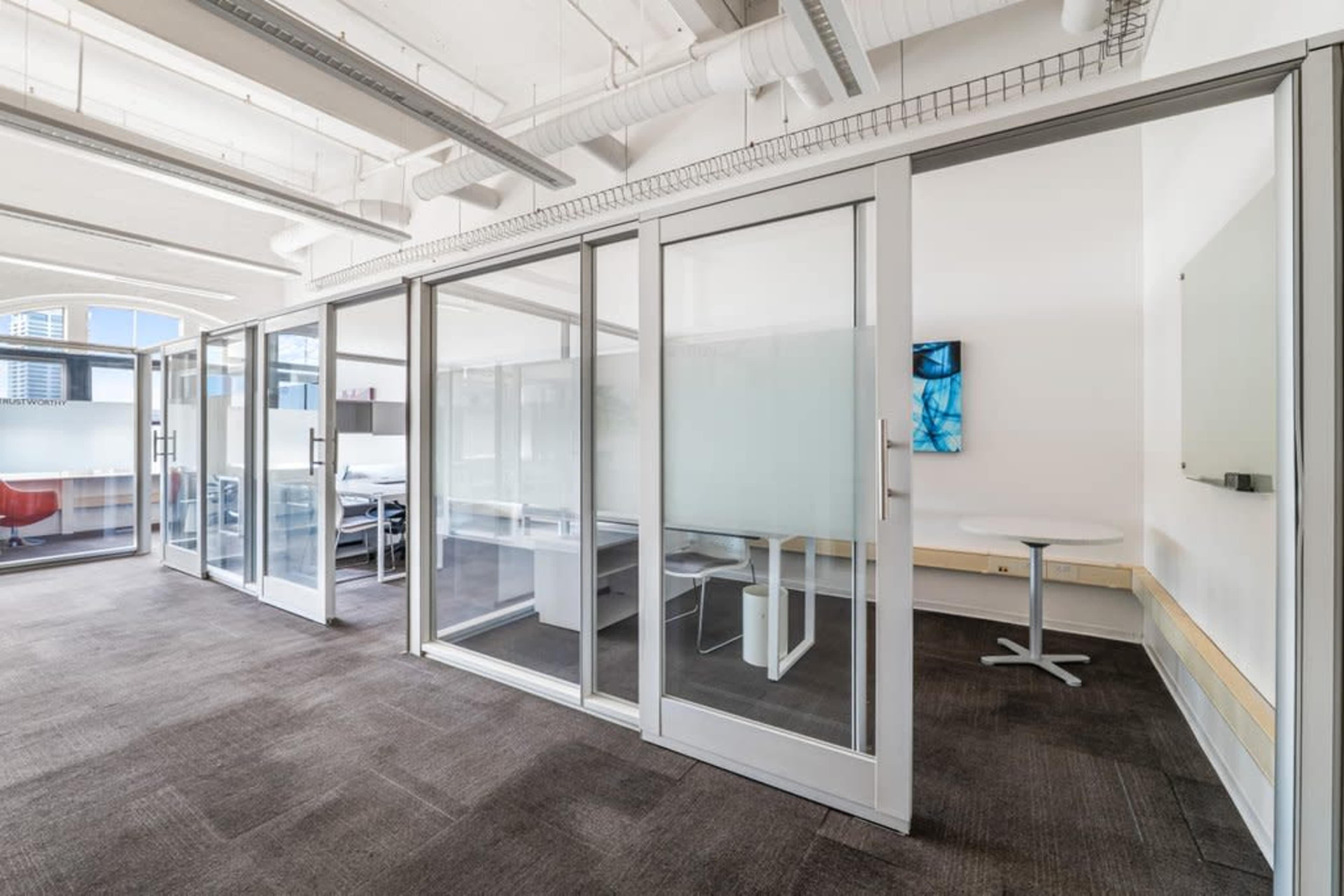 The image shows a modern office space featuring a series of glass-walled meeting rooms and a small table in the corner.