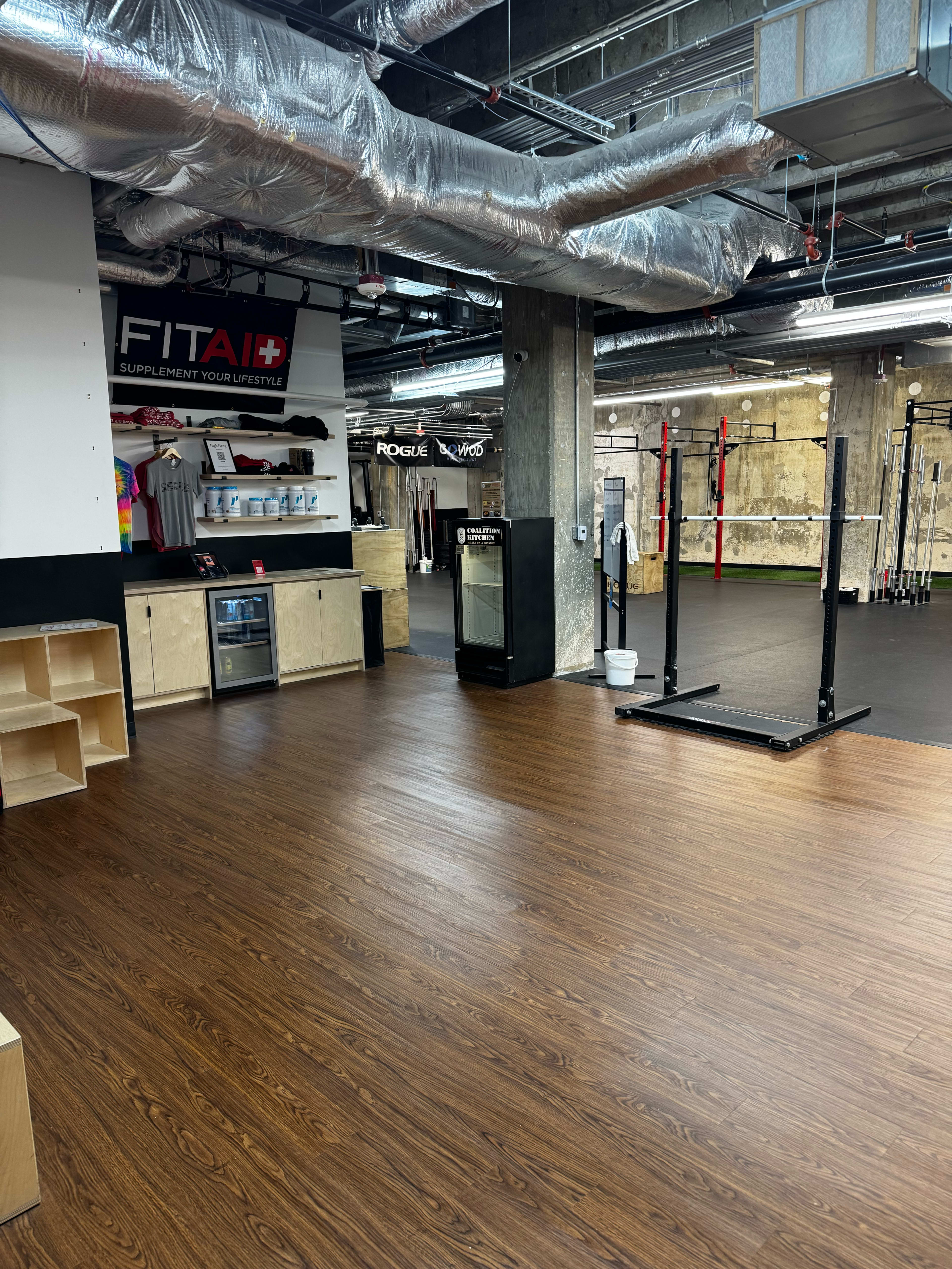 The image shows a gym interior with a wooden floor, exercise equipment in the background, and a counter displaying products.