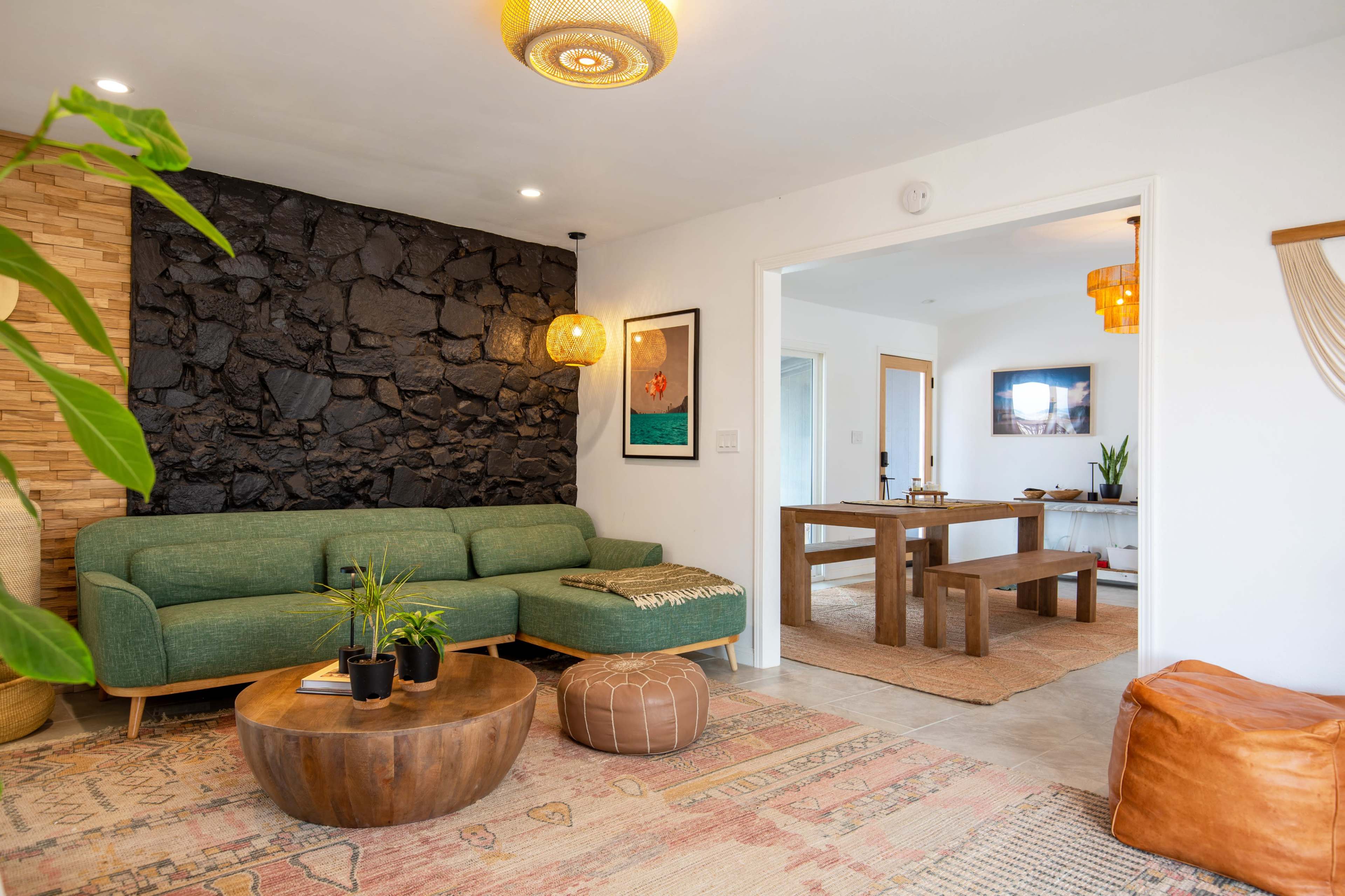 The image shows a cozy living room featuring a green sofa, a circular wooden coffee table, and a stone accent wall, with a view into a dining area beyond.