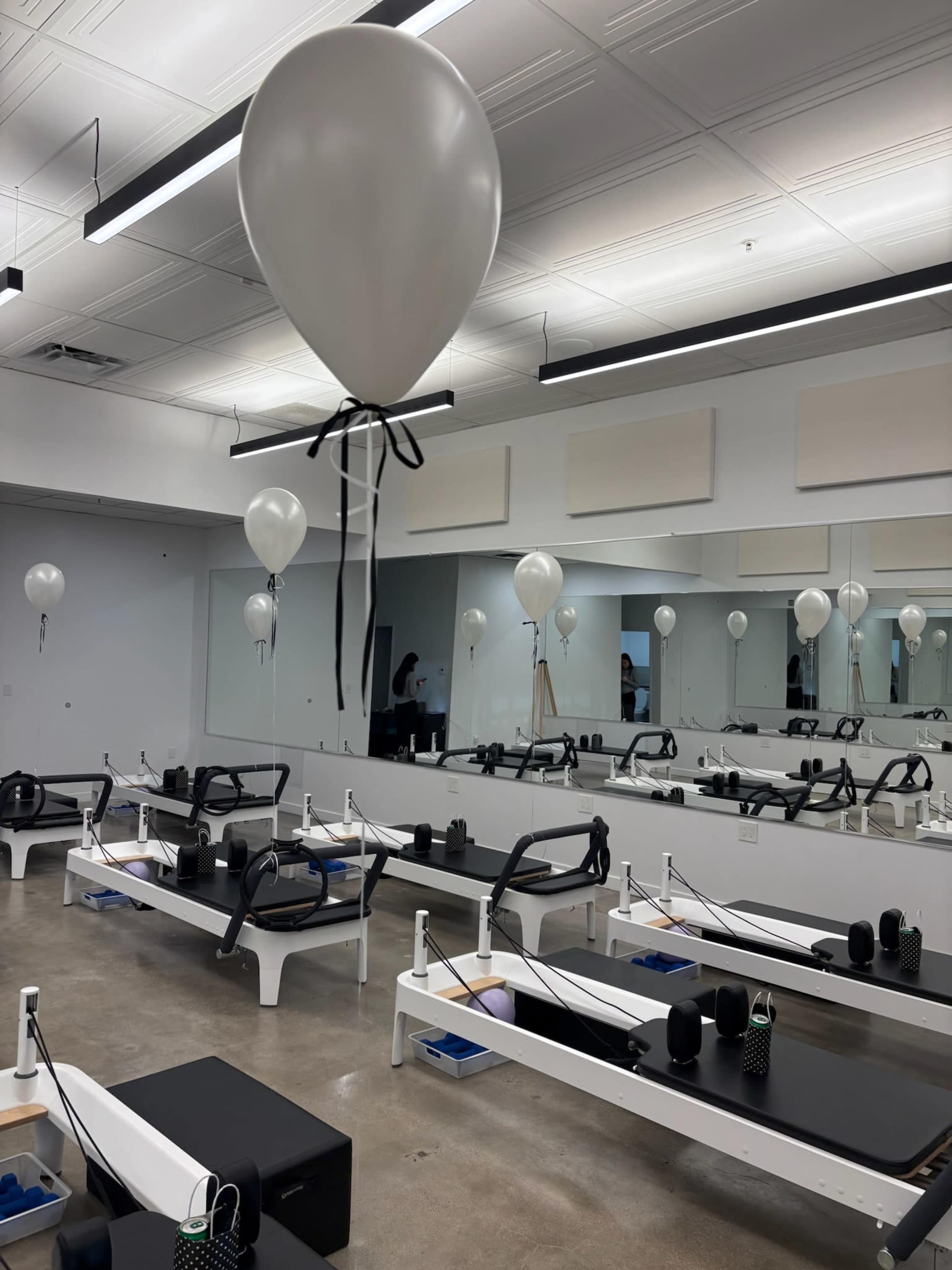 The image shows a Pilates studio with several reformer machines and white balloons tied with black ribbons hanging from the ceiling.